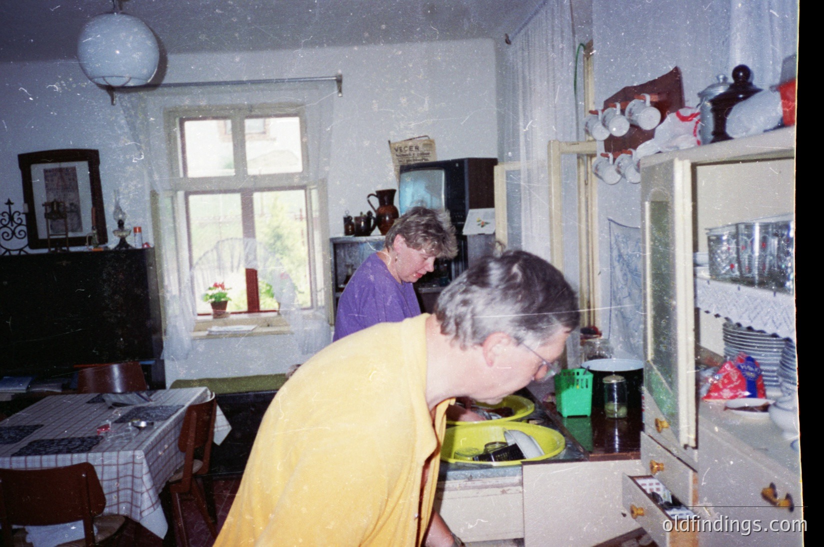 Vintage kitchen interior featuring two women in mid-20th century attire. One woman in yellow stands at a cluttered counter with a green jar, while another in purple stands near a window with floral curtains. Wooden furniture, a vintage stove, and a hanging light fixture dominate the space. Decorative items like a framed mirror and ceramic jugs add character. Likely Eastern European, mid-1900s.