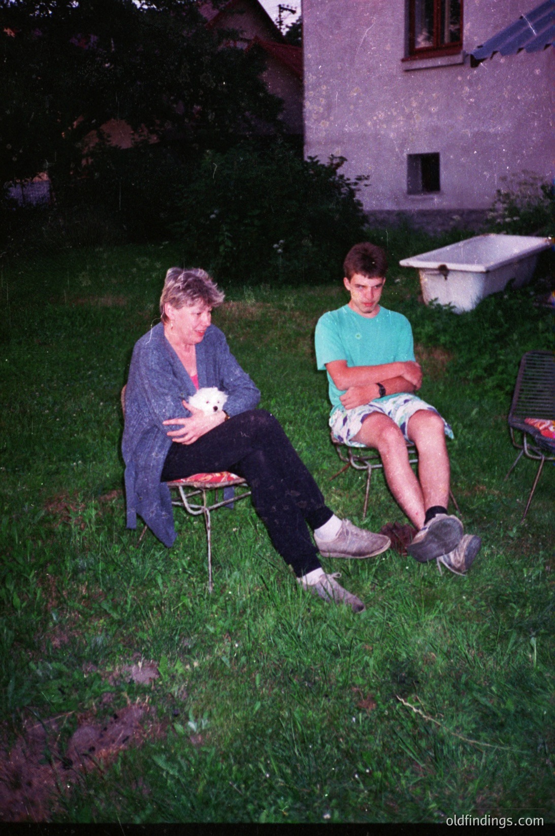 Vintage outdoor portrait of an adult woman and adolescent boy seated on metal chairs in a grassy yard. Woman wears a cardigan and dark pants, holding a small white object; boy in a teal shirt and shorts. Blurry vintage filter suggests late 20th-century photography (, ). Residential setting with a white bathtub in background (, , , ).