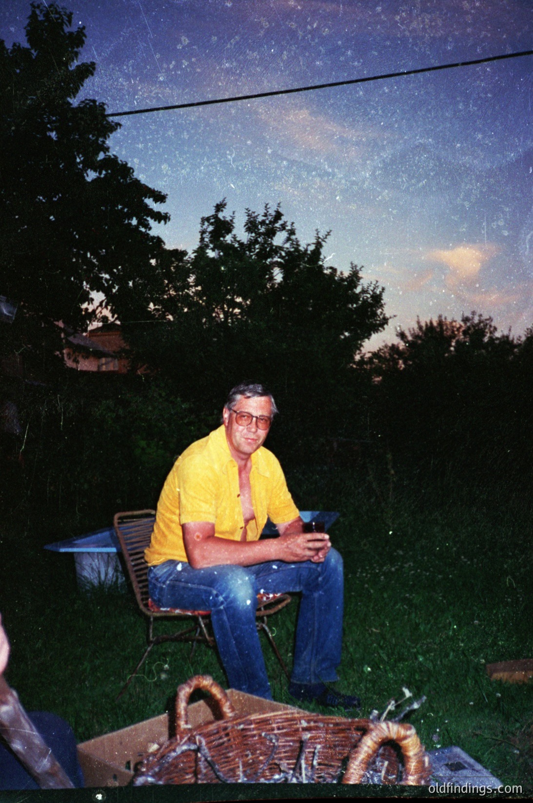 Mid-20th century man in a yellow polo shirt and glasses poses outdoors under twilight sky. Wicker chair and basket with woven handles in foreground. Lush greenery and residential rooftops visible.