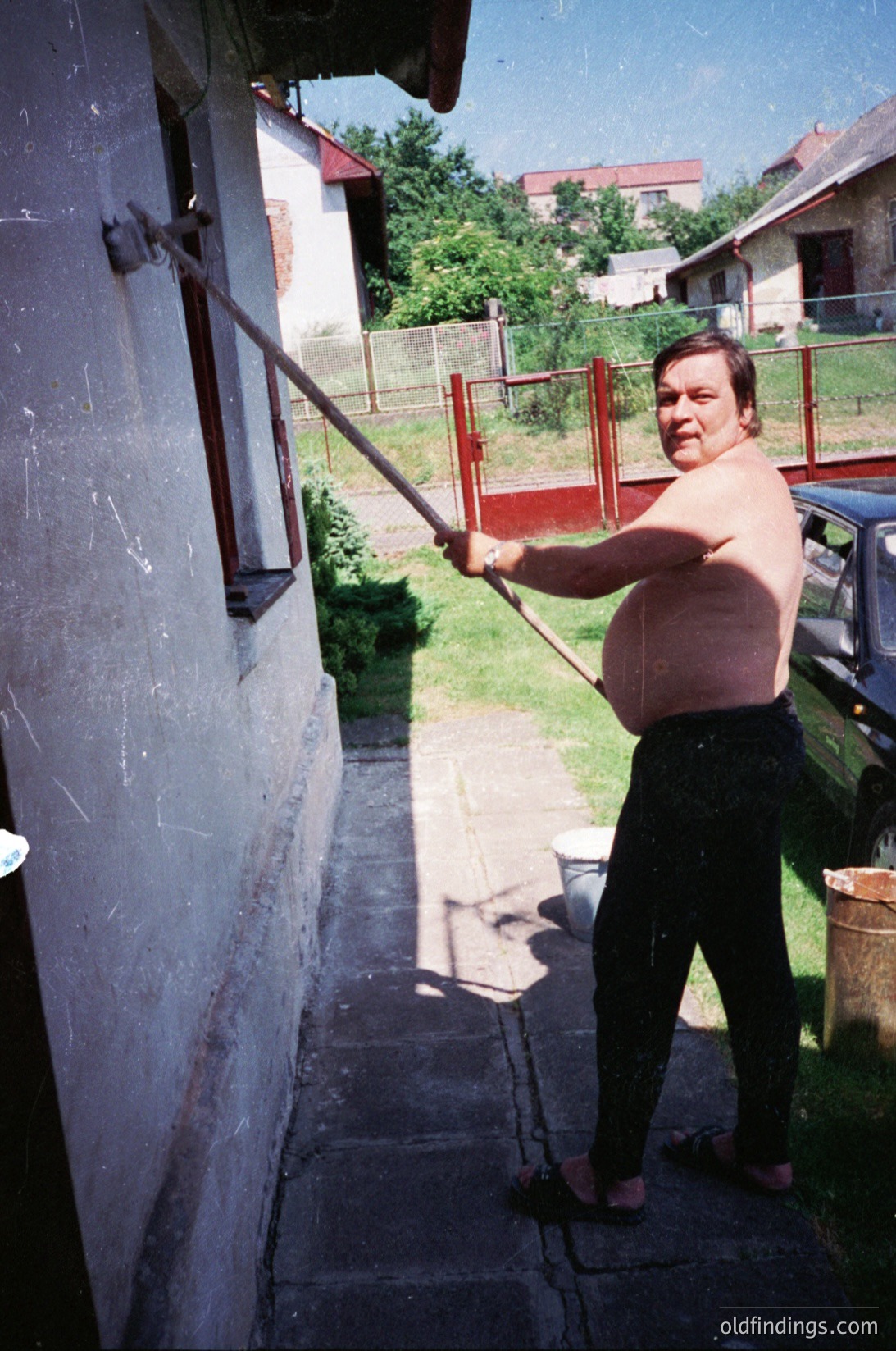 Man in mid-swing with a long wooden pole, likely a traditional *koprižnik* (pole-dancing tool), in a residential courtyard. Whitewashed walls and red fence suggest Eastern European rural architecture. Vintage car and faded colors indicate late 20th-century setting, possibly . Ideal for cultural/historical research or folk tradition documentation.