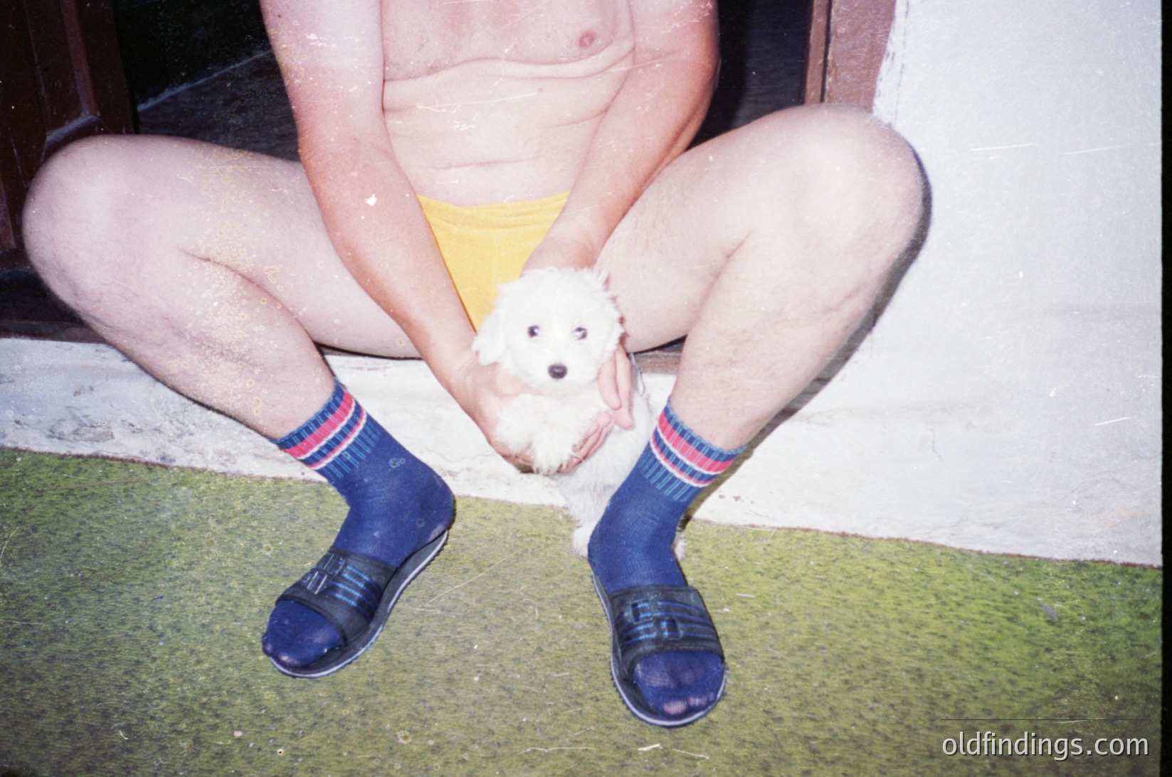 Vintage candid of a person kneeling on artificial turf, holding a small white dog. Wearing yellow shorts, blue socks with red/white stripes, and black-and-blue slip-on shoes. Indoor setting with plain wall and doorway. Evokes mid-20th century domestic life.
