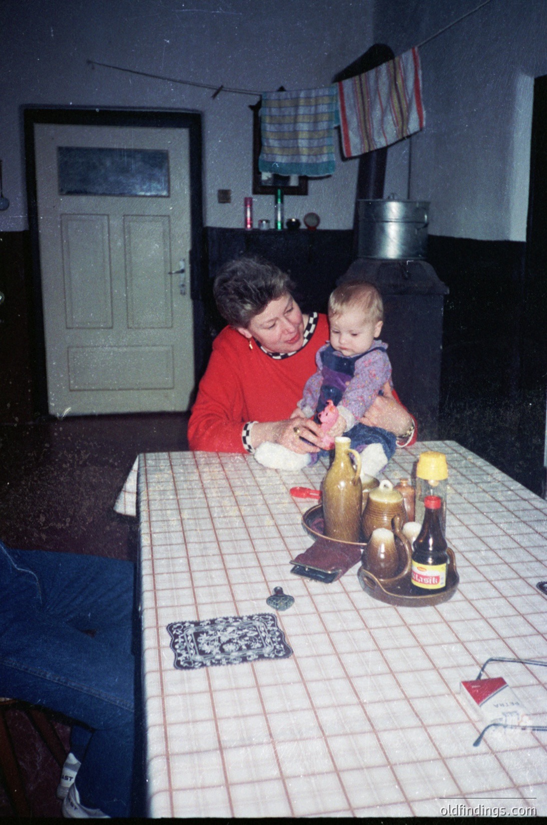 A woman in a red sweater interacts with a baby at a table covered with a checkered tablecloth, surrounded by vintage bottles and a small decorative coaster. The setting appears to be a modest indoor kitchen or dining area with a metal stove and a wooden door in the background. [Vintage family moment in a 1970s-80s kitchen, featuring generational bonding over simple objects like bottles and a checkered tablecloth ]