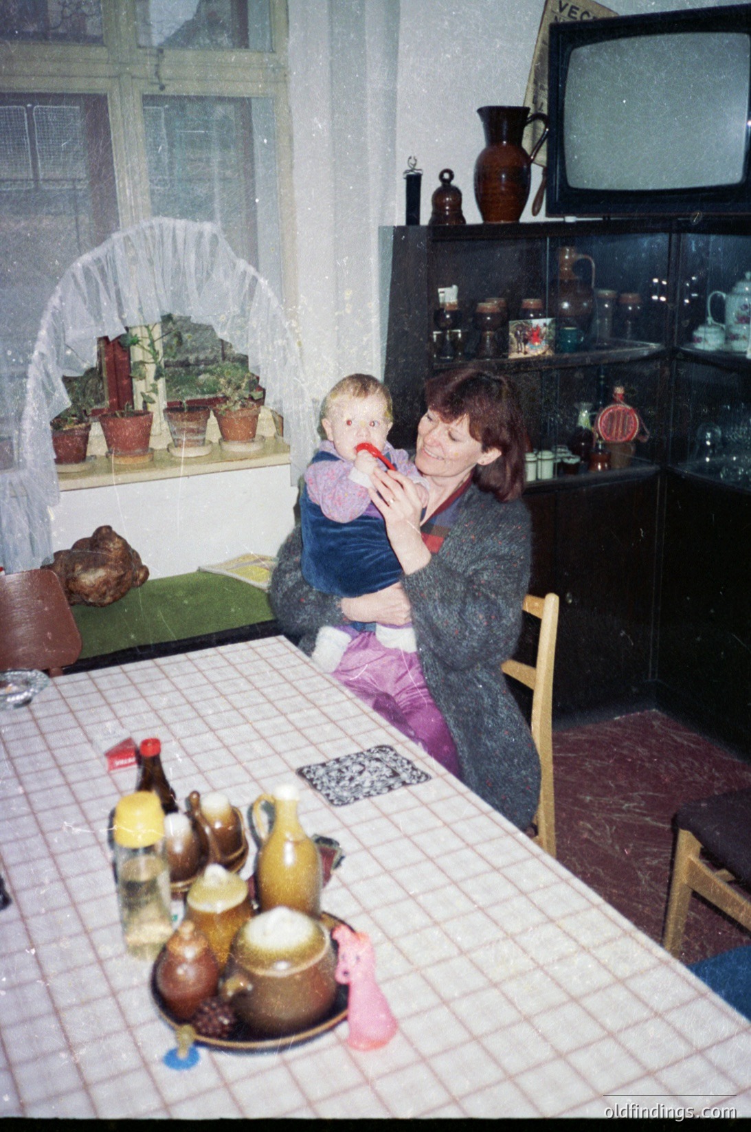 A woman cradles a young child indoors, likely mid-1980s–90s Eastern Europe. The scene captures a modest kitchen with a tiled table displaying ceramic jars, a pitcher, and a teapot. A small TV sits atop a wooden cabinet filled with pottery. A fireplace alcove with potted plants and a woven arch adds warmth. The setting reflects everyday domestic life during the Soviet-era or post-communist period.