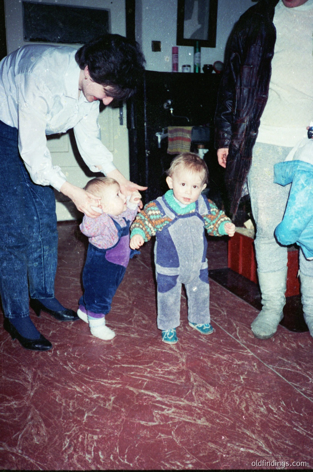 Two toddlers learning to walk indoors, guided by adults in a 1990s-style home. Bright red vinyl flooring contrasts with patterned children’s overalls and adult jeans. Kitchen appliances and a coat rack in background suggest domestic setting.