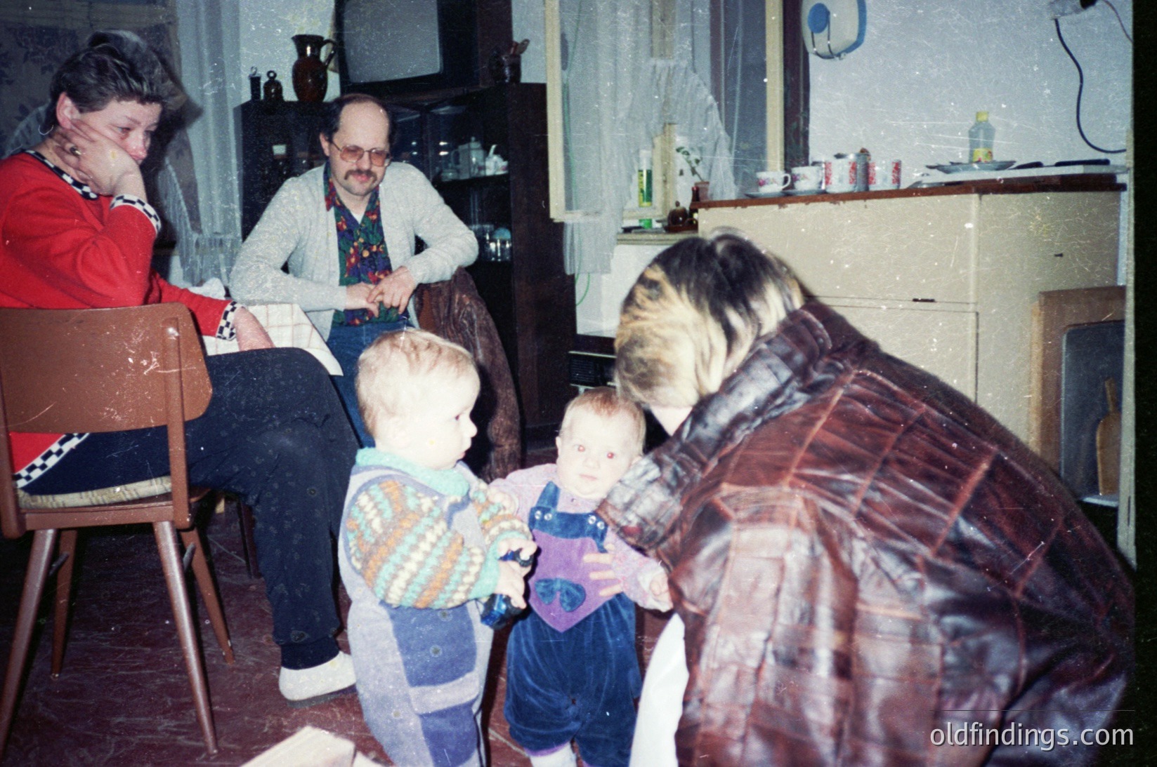 Vintage indoor family moment: adults seated on worn furniture, two young children playing on a linoleum floor. Kitchen cabinets and a stove in background suggest a mid-century domestic setting. Colorful clothing and patterned blankets hint at 1970s-80s Eastern European style.