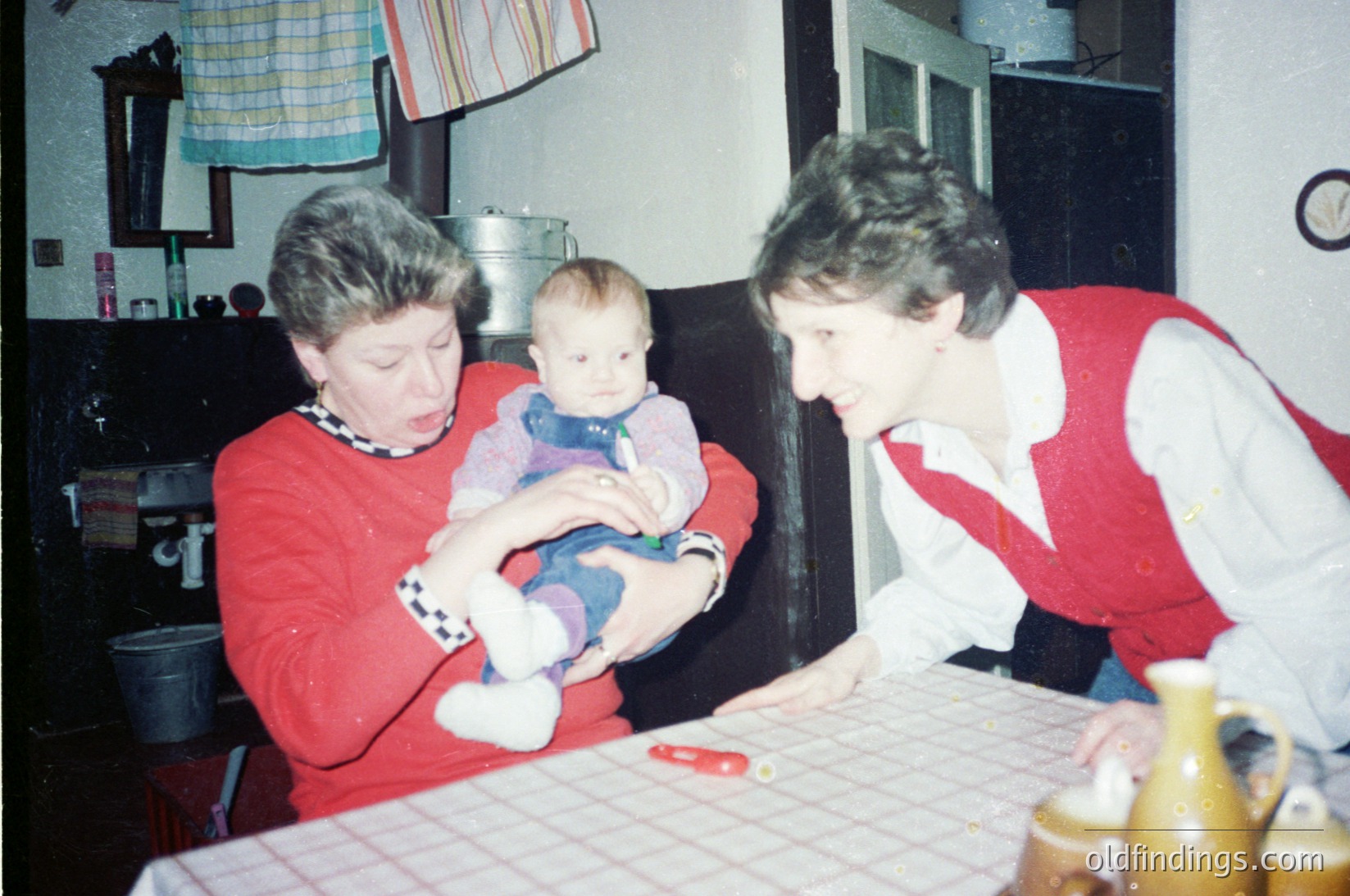 Two women in 1970s-style red sweaters share a moment with a baby in a modest kitchen. Checkered curtains and a tablecloth with floral patterns suggest a mid-century domestic setting. A knife rests on the table, hinting at meal prep.