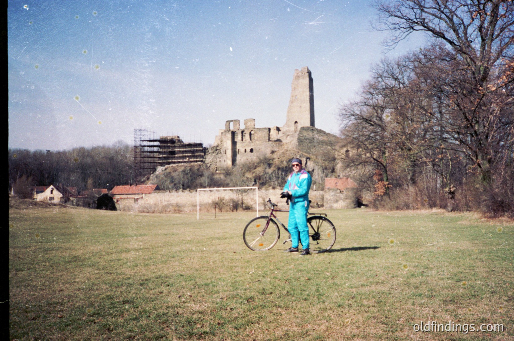 Vintage photo of medieval ruins with a lone cyclist in bright teal cycling gear. Crumbling stone tower and walls in a grassy field, surrounded by sparse trees. Likely Eastern Europe, 1970s-1980s.