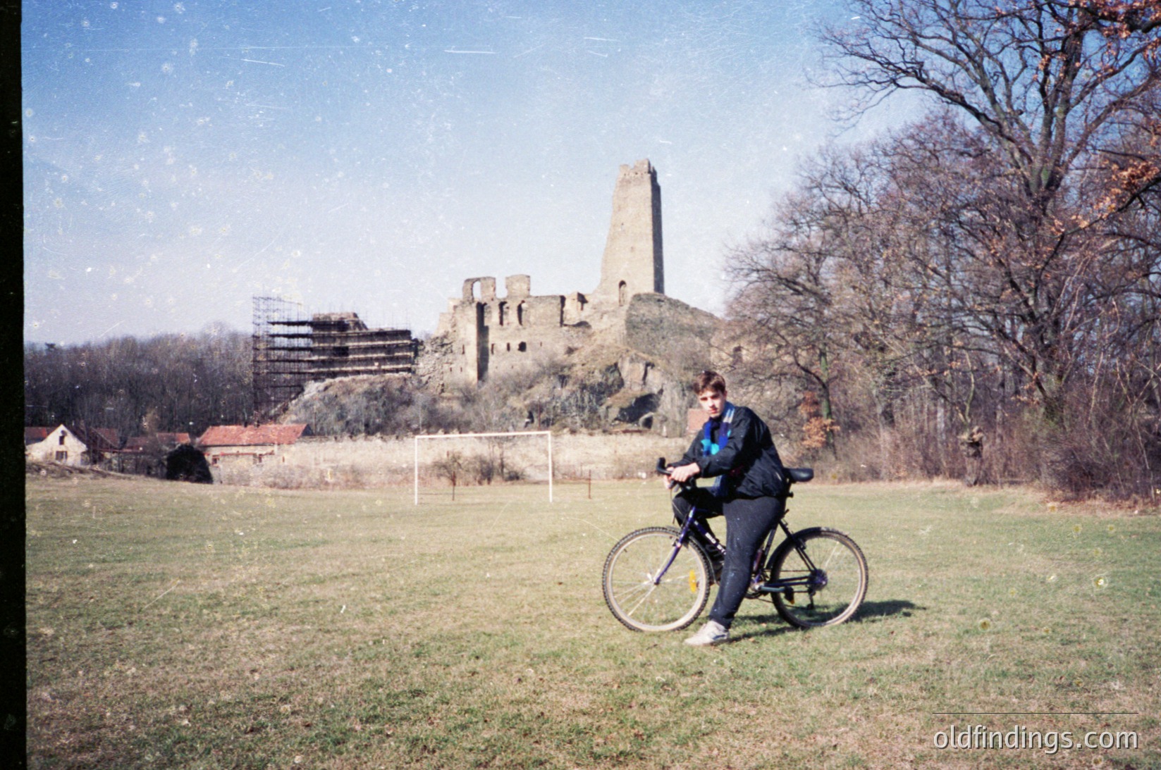 Vintage photo of a person in cycling attire riding a basic frame bike near a medieval stone ruin with a tower and arched windows. Open grassy field with sparse trees and a fence in background. Likely Eastern Europe, 1980s-1990s.