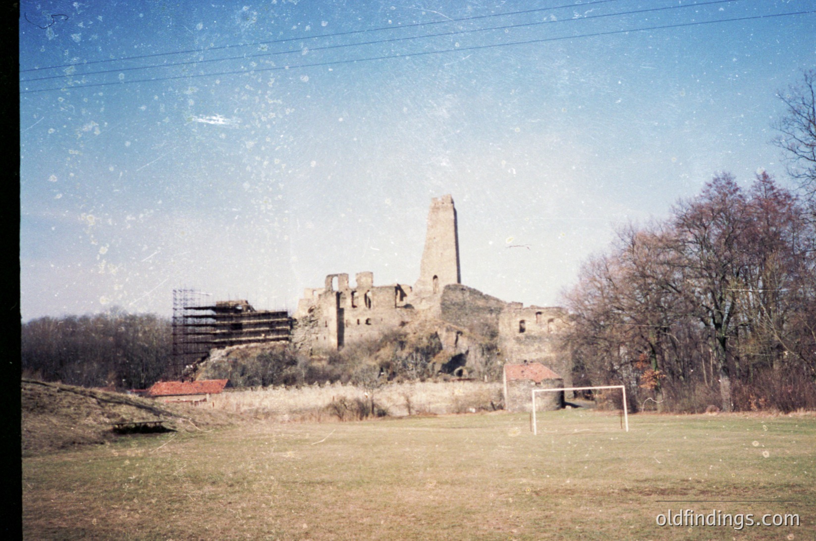 Vintage photo of a ruined medieval tower and stone walls, likely Eastern European. Overgrown grass and a soccer goal in foreground suggest repurposed land. Scaffolding indicates partial restoration. *(Note: Time period inferred from color tone and film grain; location speculative but plausible for Eastern European ruins.)*