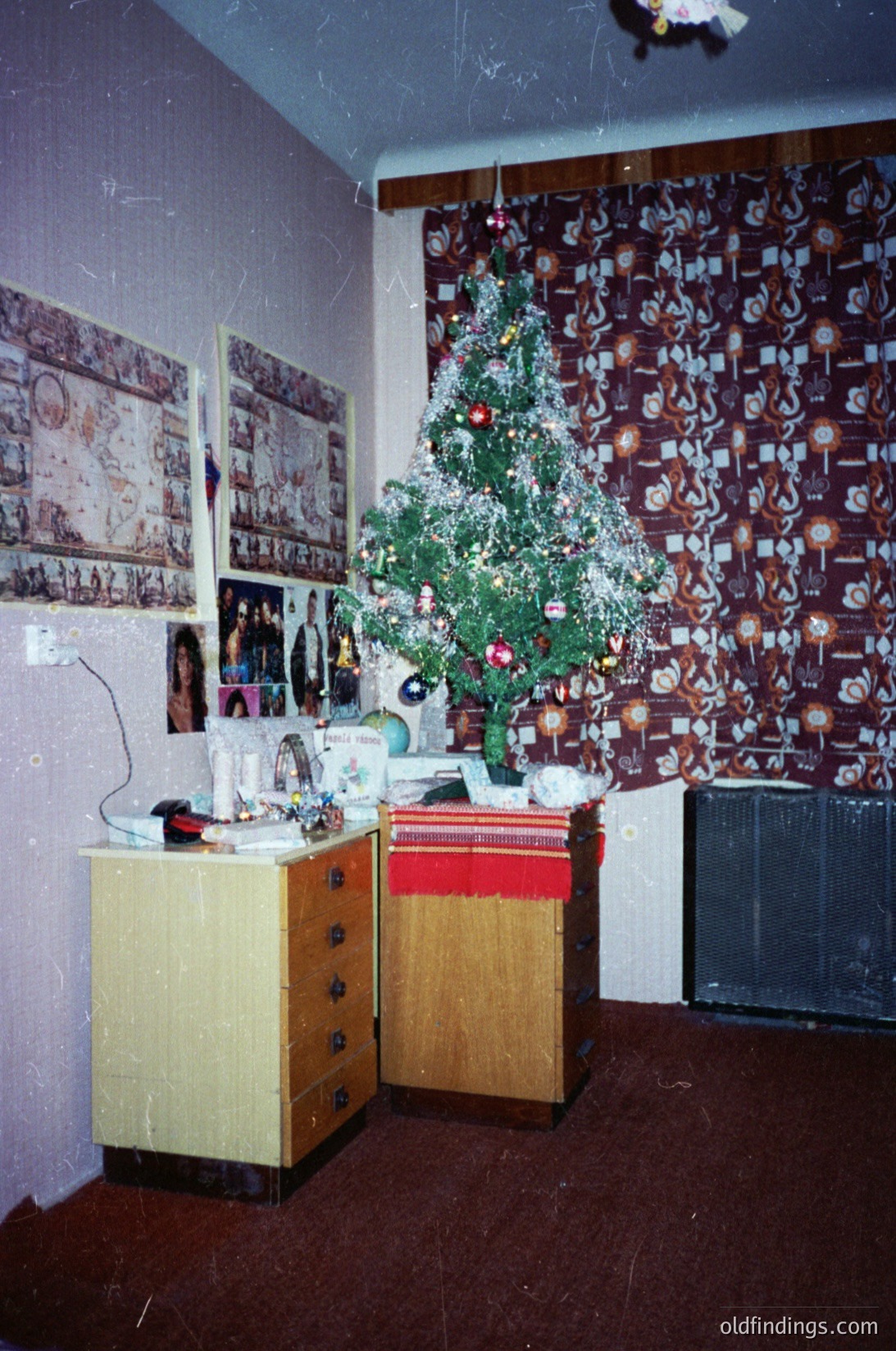 Vintage indoor scene featuring a modestly decorated Christmas tree with tinsel and ornaments against a wall adorned with framed photographs and Soviet-era propaganda posters. Wooden dressers with drawers and a vintage radio/tape player occupy the foreground. Warm, aged lighting suggests a 1970s–1980s Eastern Bloc home setting.