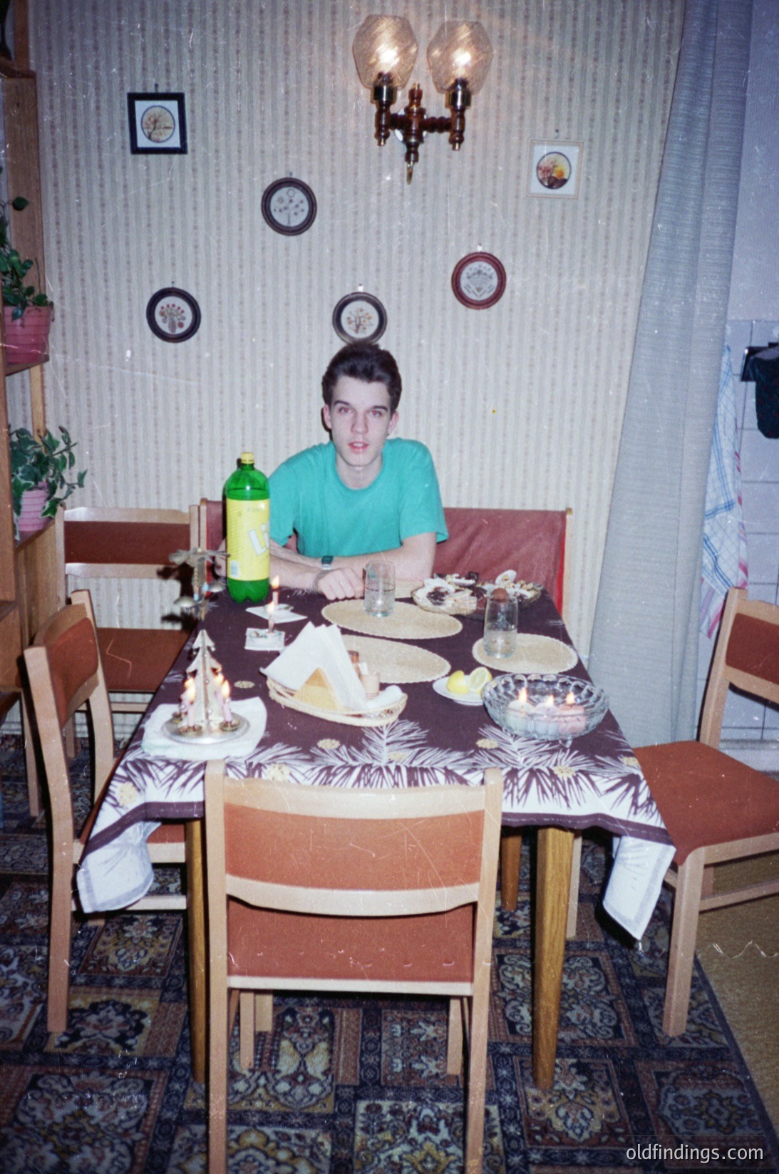 Vintage dining scene featuring a young man seated at a wooden table draped in a patterned tablecloth, surrounded by mismatched chairs. Decor includes wall-mounted round plaques, a chandelier, and potted plants. Glassware, a bottle of yellow liquid, and a bowl of food are on the table. Likely Eastern European home, 1980s–1990s.