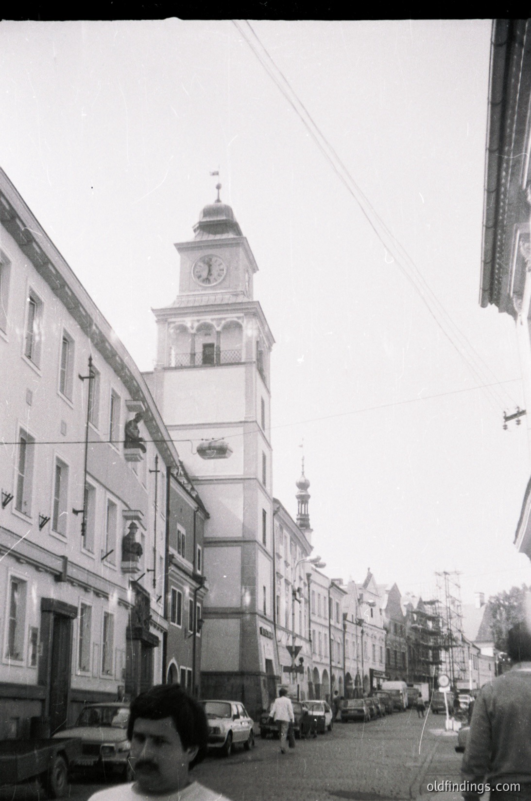 Prominent clock tower with ornate dome and Soviet-era architectural details dominates a narrow European street. Mid-20th century urban scene with parked vintage cars and pedestrians. Likely Warsaw, Poland,