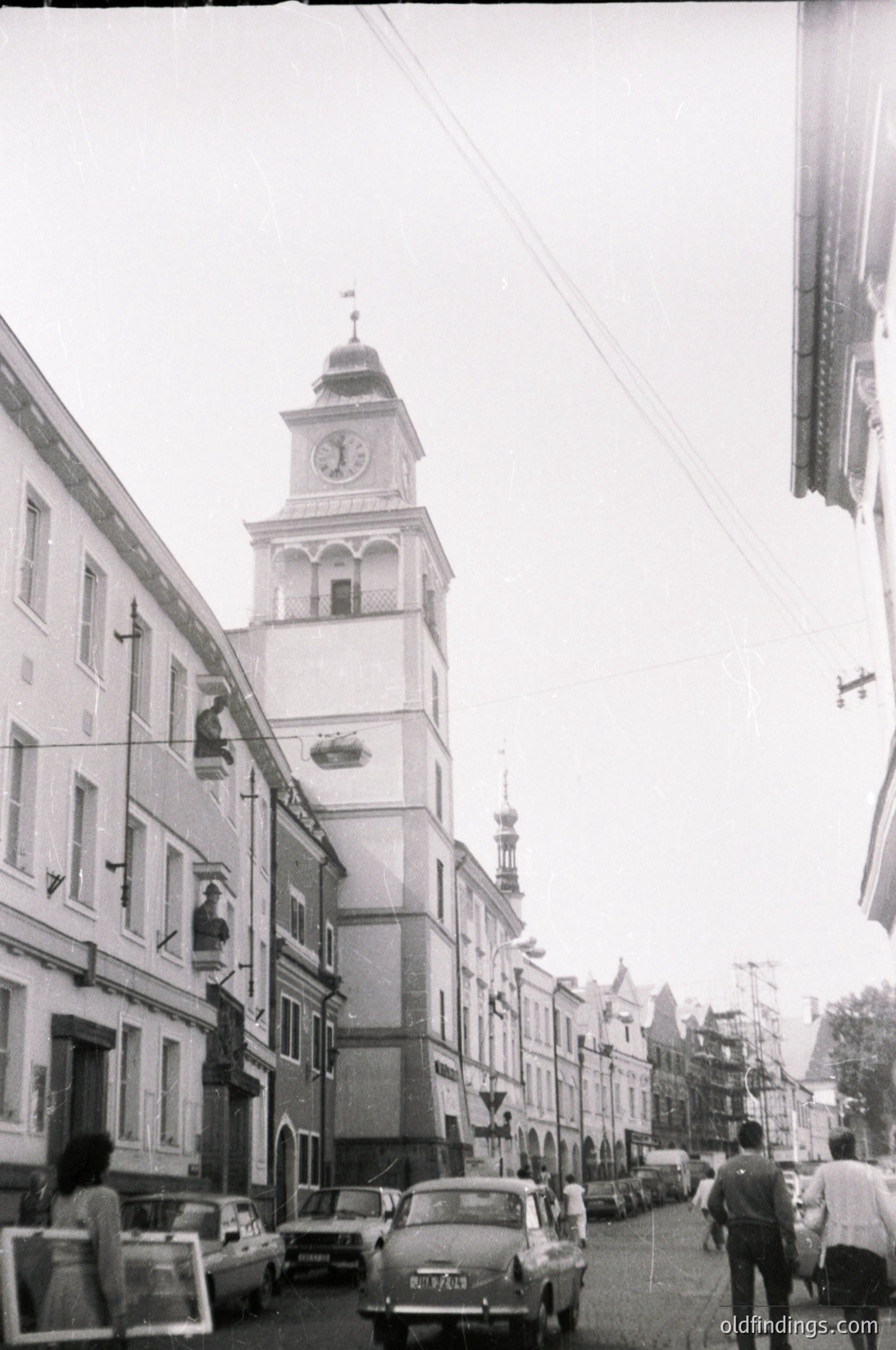Black-and-white street scene featuring a prominent **clock tower** with a domed roof, flanked by **1960s-era buildings** in a European urban layout. **Vintage cars** (including a white sedan) line the street, and pedestrians walk along the sidewalk. Overhead wires and minimal greenery suggest a mid-century urban environment.