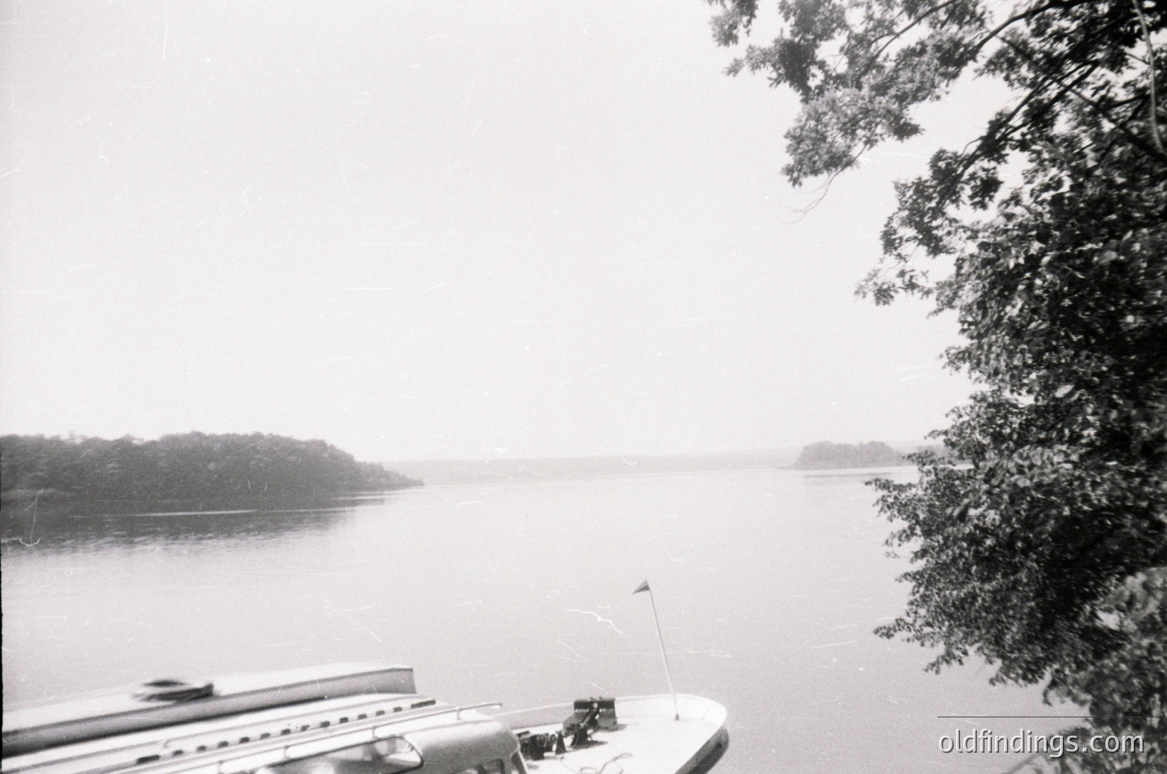 Mid-20th century black-and-white lakeside scene with misty horizon. A docked boat’s edge and flagpole frame the shot, while a distant train crosses a bridge on the left. Dense foliage partially obscures the top right corner.
