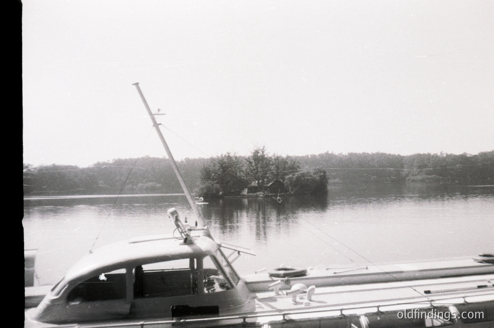Mid-20th century boat docked at a serene waterfront, likely a lake or coastal inlet. The vessel features a tall mast with a flagpole, suggesting recreational or small-scale commercial use. In the background, a small island or landmass with scattered structures and greenery. Overcast skies enhance the vintage, monochrome aesthetic.