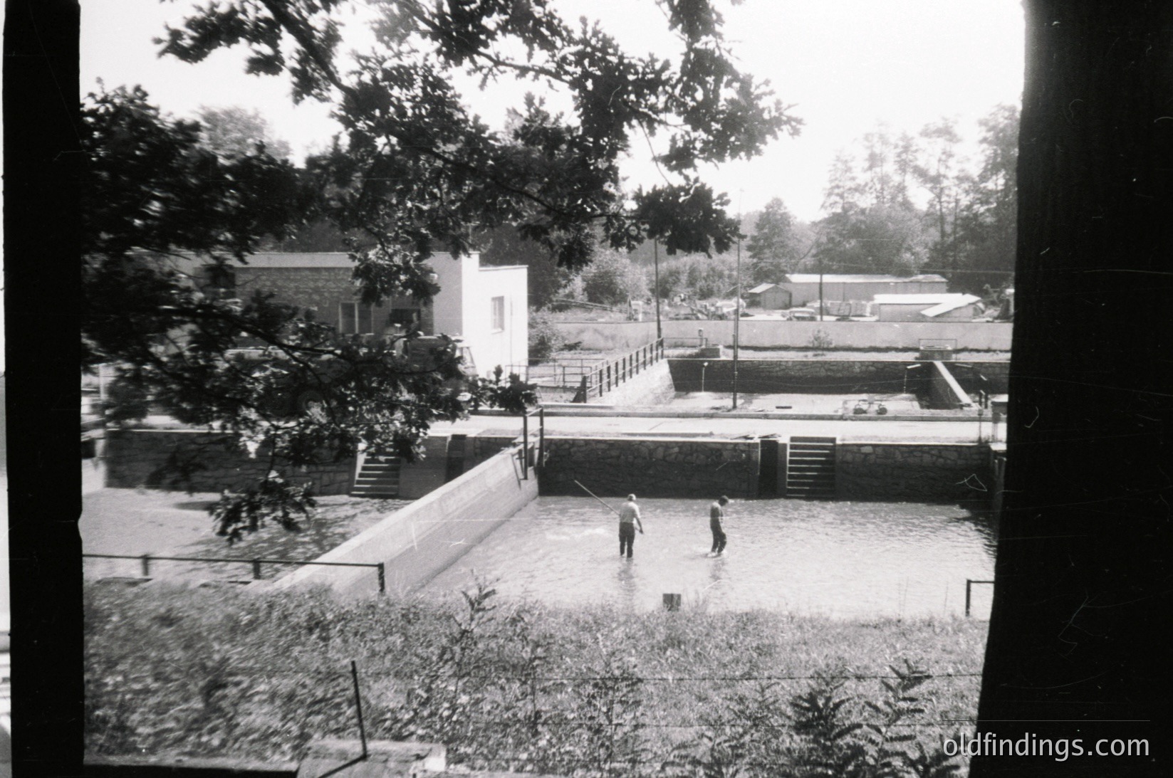 Black-and-white snapshot of a flooded residential courtyard, likely mid-20th century. Two children wade through shallow water near a concrete pathway, framed by overgrown vegetation and a white building with a flat roof. Rainwater pools around a brick structure in the background.