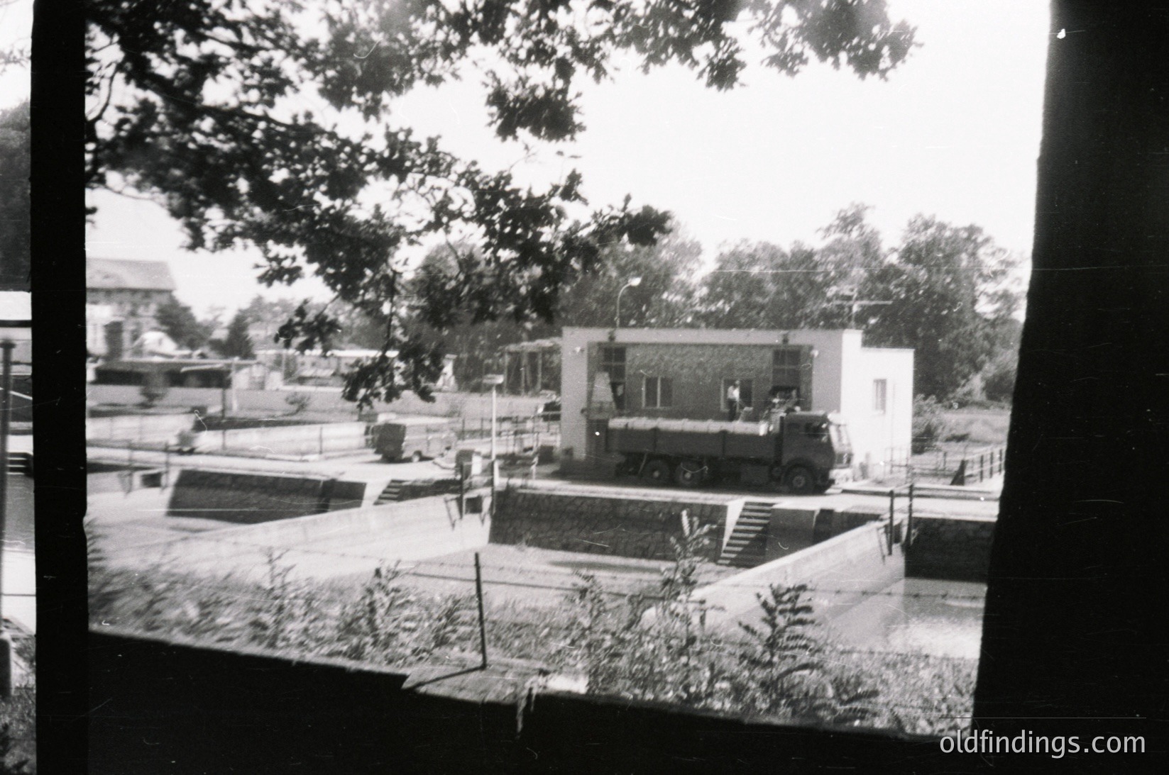 Vintage black-and-white photo of a mid-20th century courtyard featuring a low-rise, modernist building with flat roof and minimalist design. A military truck with mounted weaponry occupies the center, suggesting Cold War-era military presence. Overgrown vegetation and a single tree frame the scene, hinting at neglect or wartime conditions. Urban setting with visible infrastructure like roads and fencing.