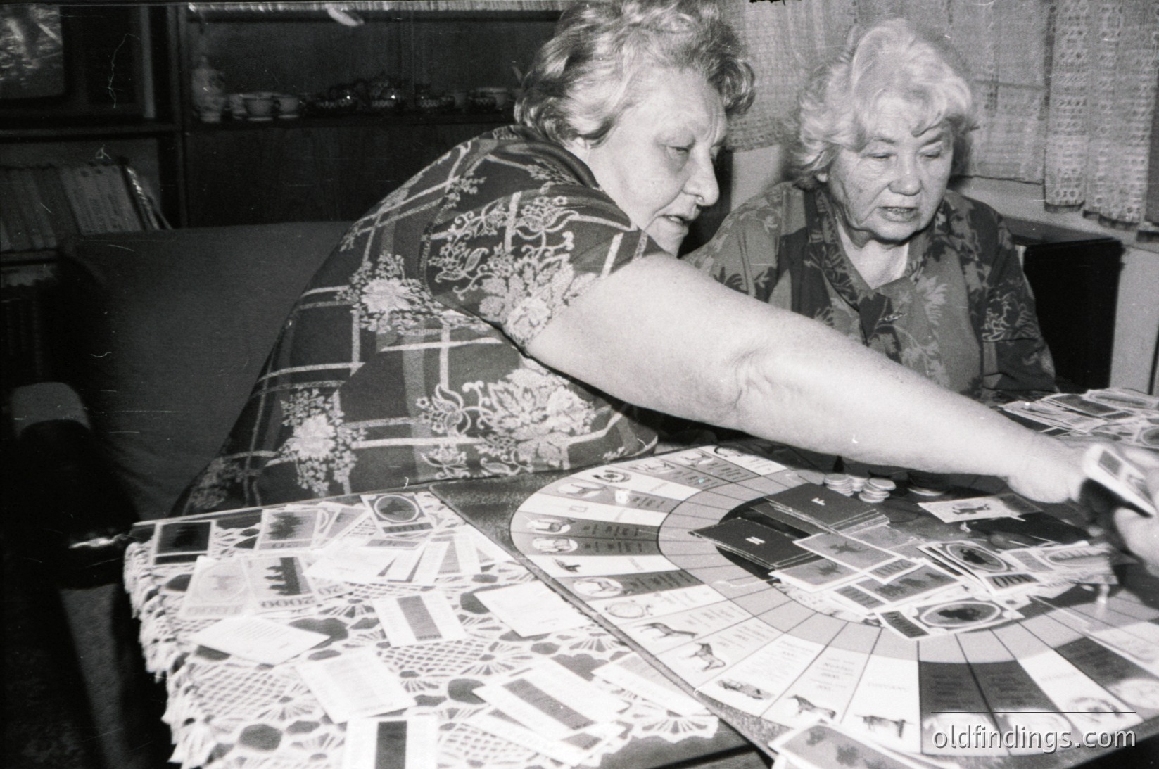 Two elderly women in mid-20th century attire (1960s–1970s) focus on assembling a jigsaw puzzle on a patterned tablecloth. The setting appears domestic, likely a living room or kitchen. Their expressions suggest concentration and camaraderie.