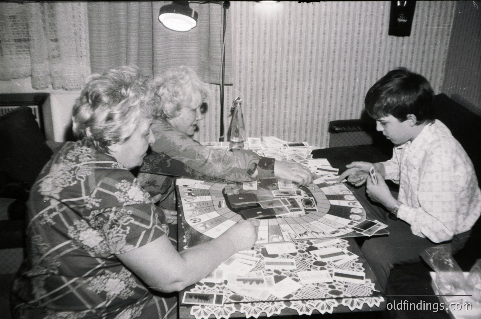 A mid-20th century indoor scene showing two adults and a child engaged in crafting. Spread across a table are paper cutouts, scissors, and a lamp with a shade. The woman’s patterned blouse and the child’s button-down shirt suggest a 1960s–1970s setting. Likely a family project or educational activity.