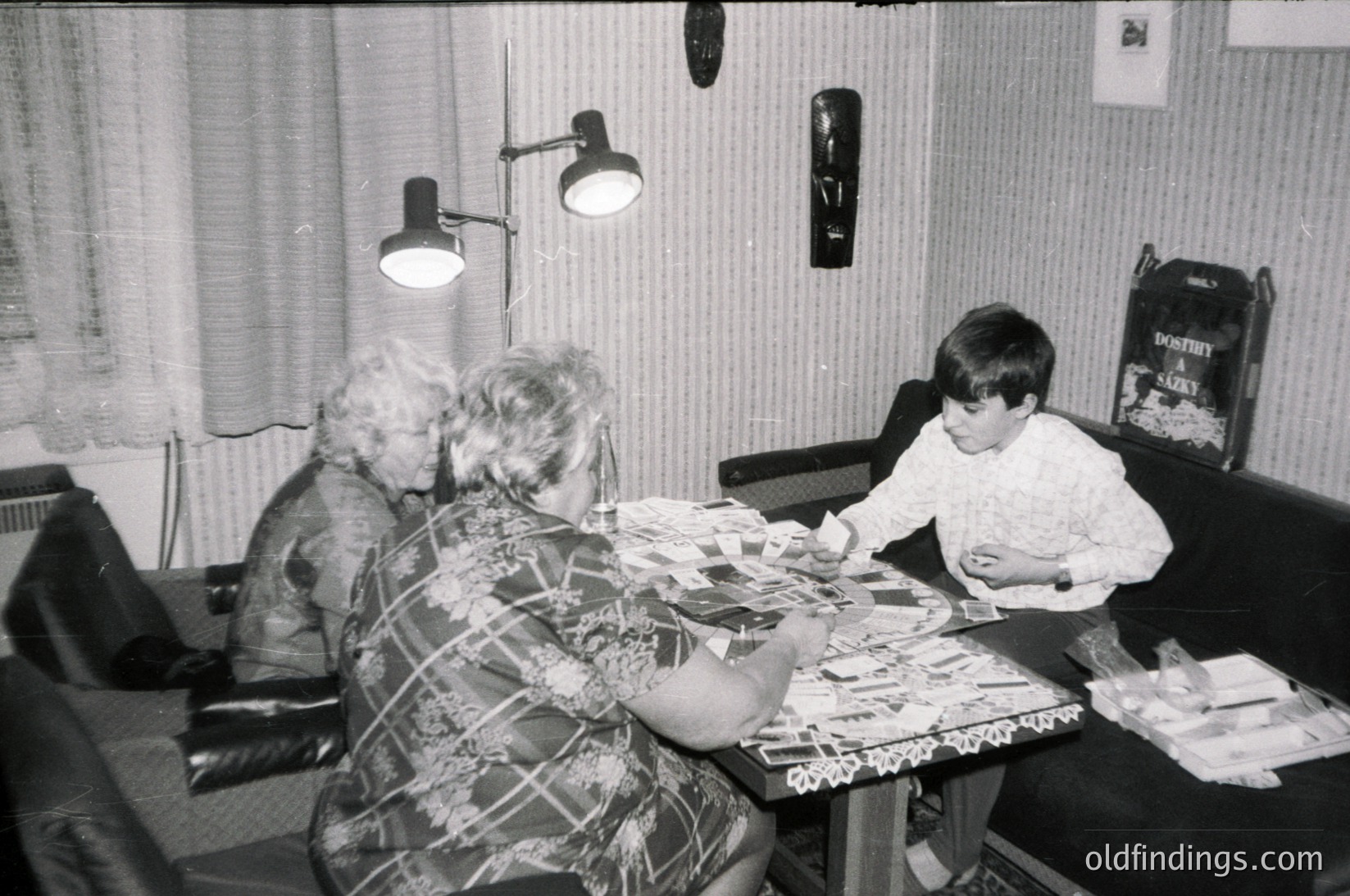 Black-and-white interior shot of three generations engaged in a craft activity, likely mid-1970s. Two women and a child sit at a cluttered table with paper, scissors, and glue. Decorative wall lamp and framed posters add vintage charm.
