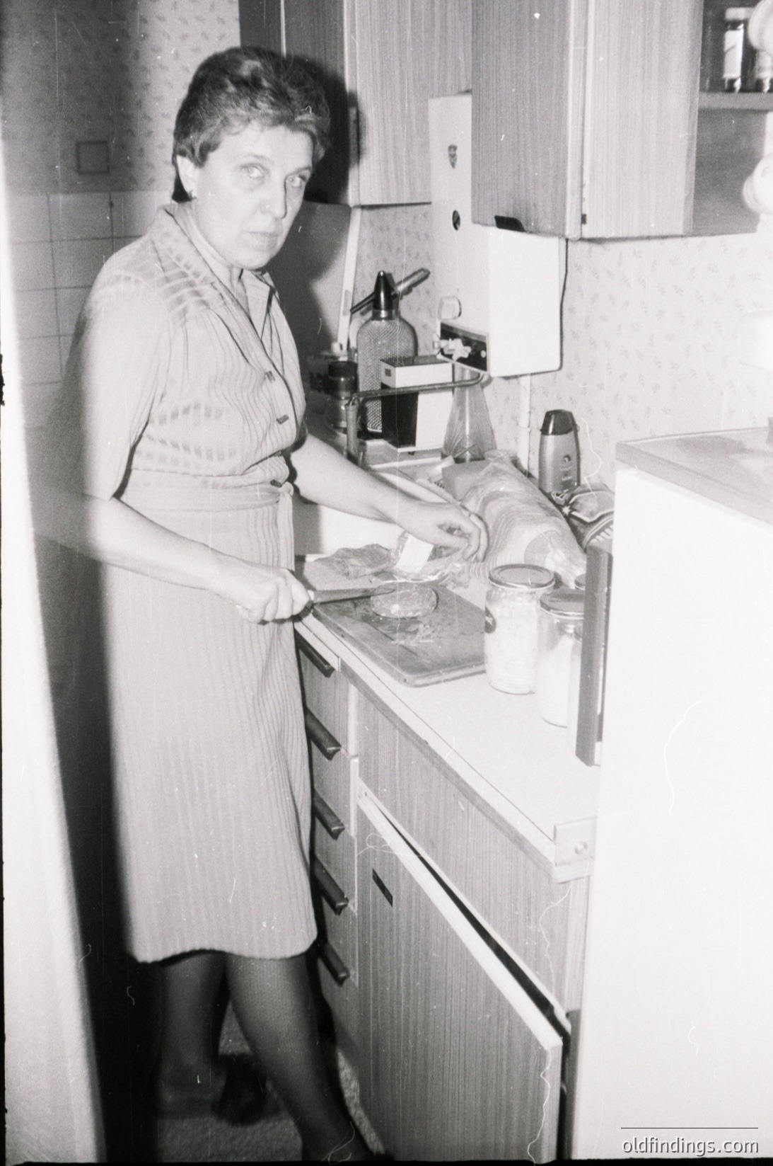 Mid-century kitchen scene featuring a woman in a patterned apron preparing food on a vintage countertop with a manual coffee grinder, canned goods, and a stove. Wooden cabinetry and a white refrigerator reflect 1960s domestic design.