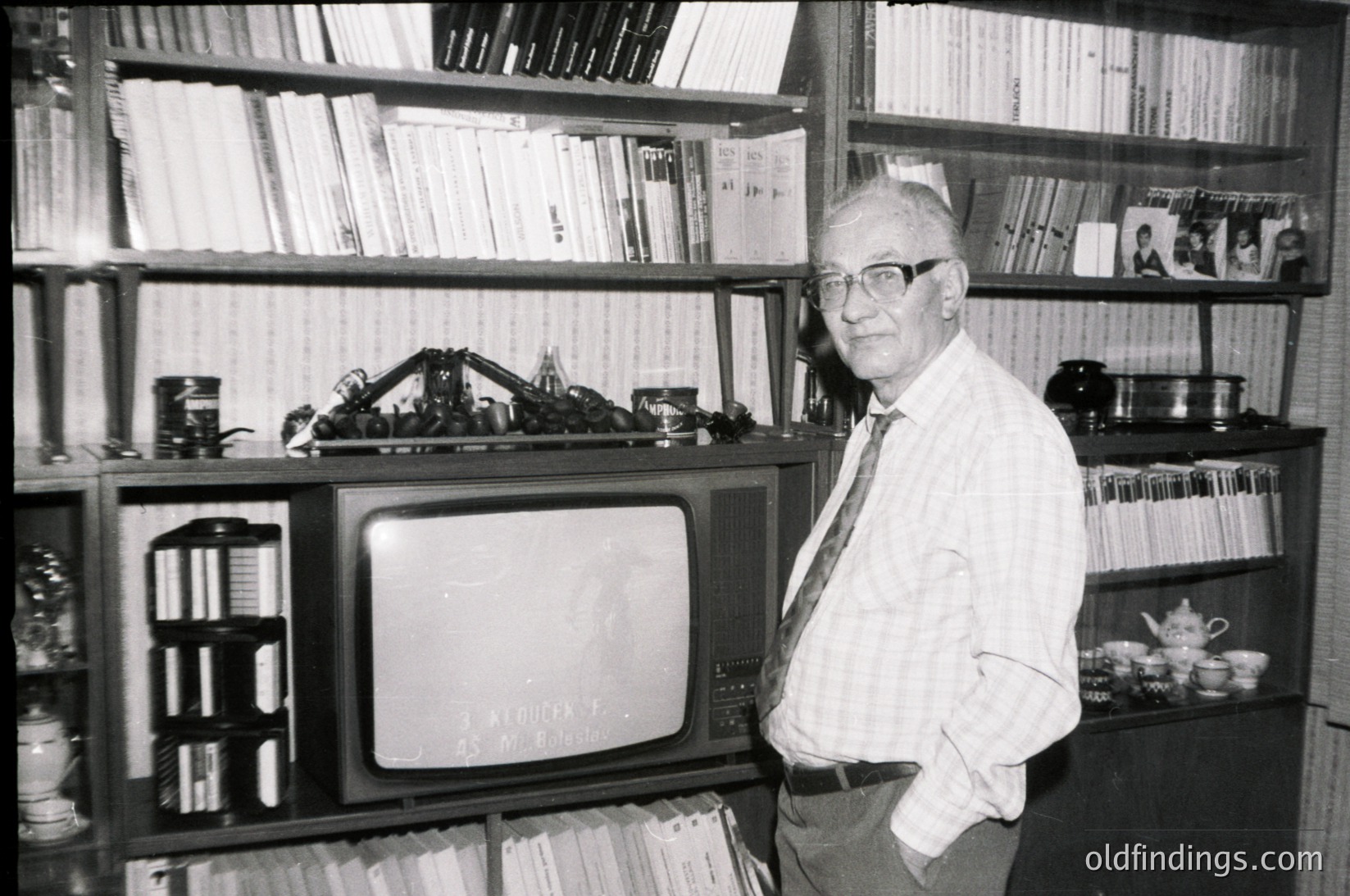 Mid-20th century man in a bookstore/library setting, posing beside a vintage CRT TV displaying static. Surrounding shelves hold books, records, and vintage cameras. Formal attire suggests professional or academic context, likely –.