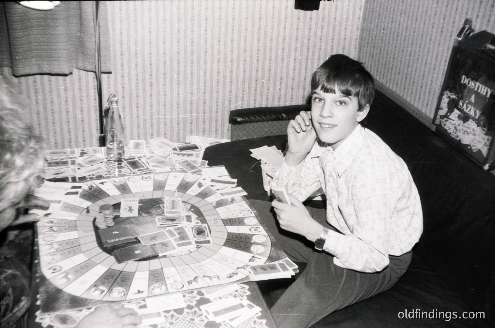 Young boy in 1970s-era home playing board game with circular layout, surrounded by scattered game pieces. Mid-century interior with vinyl upholstery, lamp, and framed poster of Dostoyevsky’s *Crime and Punishment*. Candid, informal setting captures playful engagement.
