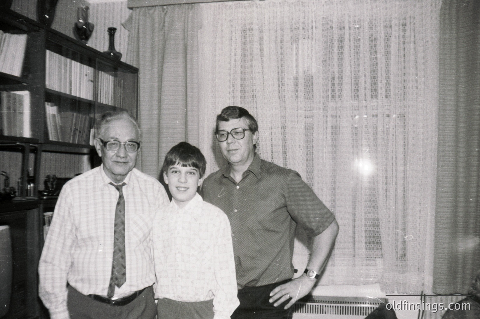 Three individuals pose indoors in a mid-century setting: an older man in a striped tie, a young boy in a button-down, and another man in a collared shirt. Bookshelves with ceramic vases and framed art fill the background. Lighting suggests evening or indoor photography.