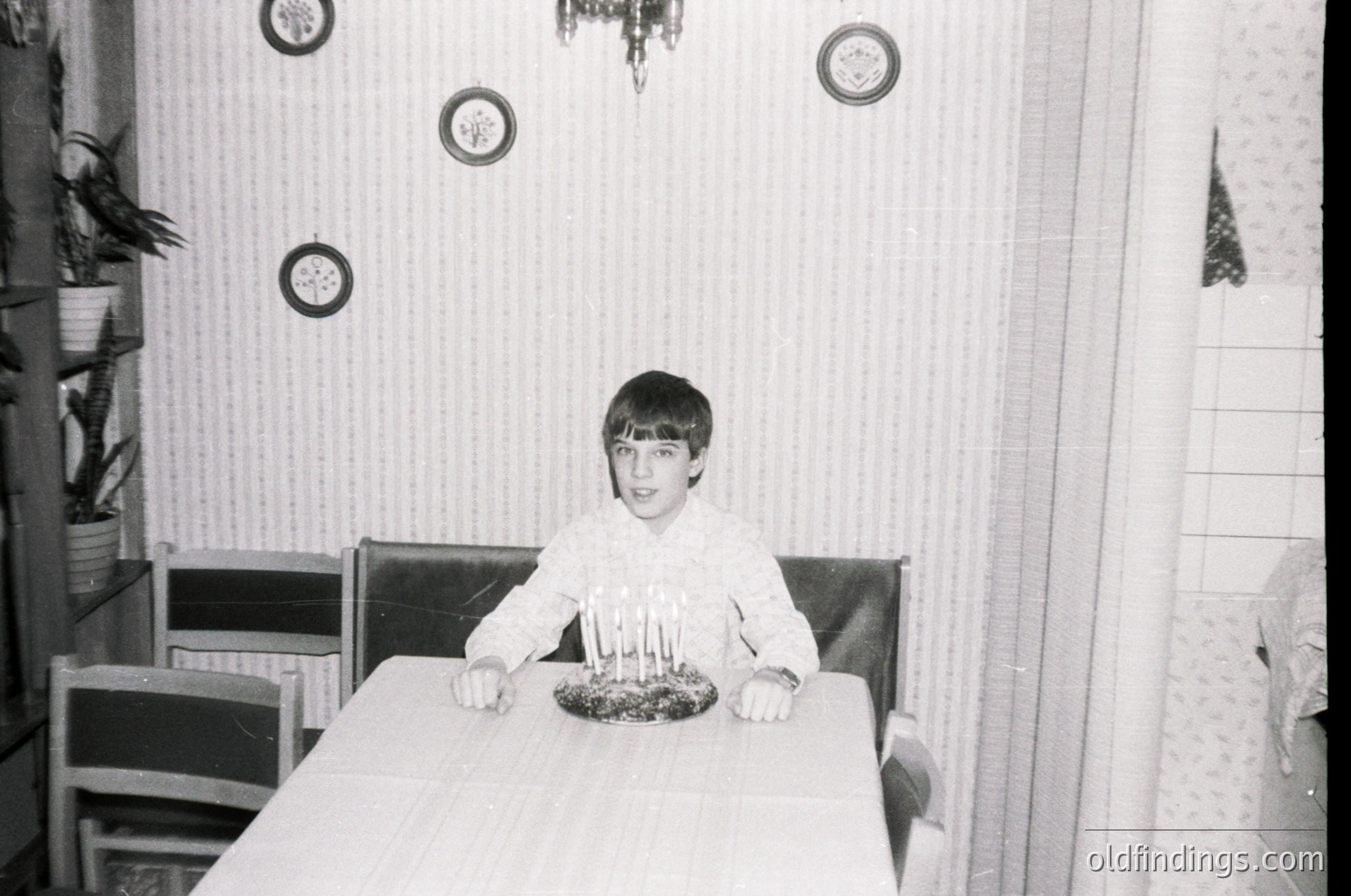 Mid-century kitchen interior with vertical striped wallpaper and round wall sconces. Young boy seated at a rectangular table, holding a birthday cake with lit candles. Wooden highchair and vintage-style chairs visible. Likely 1960s–1970s domestic setting.
