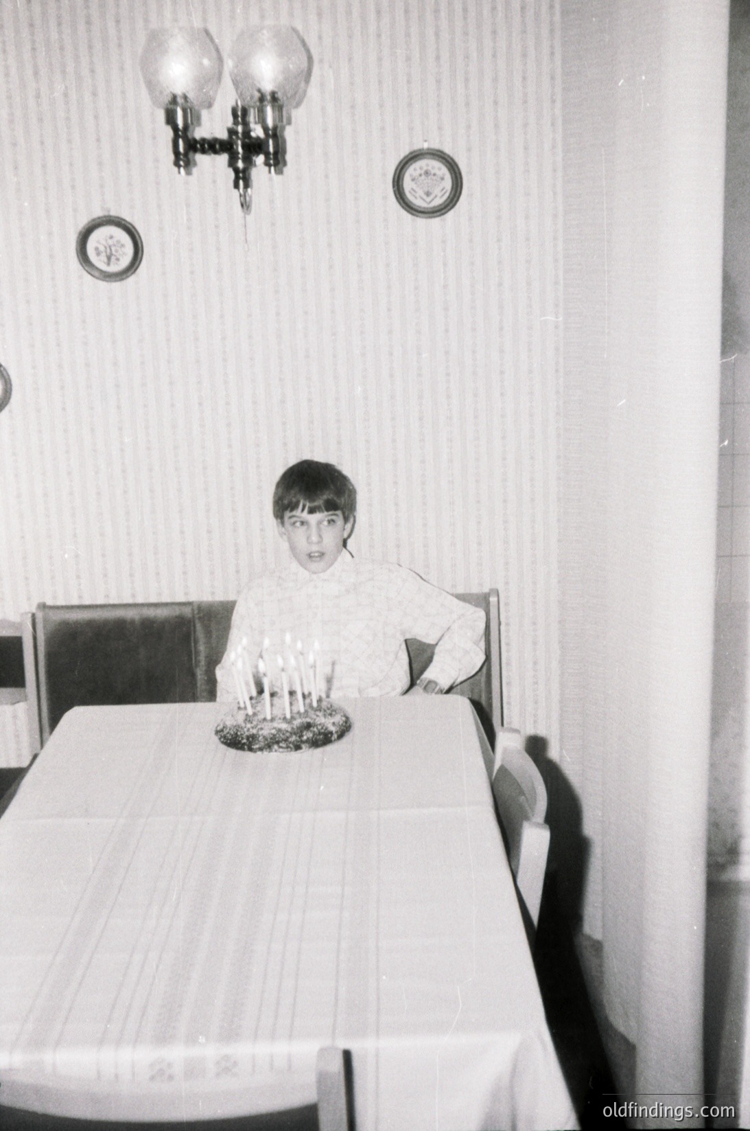 Mid-century dining scene with a young girl seated at a rectangular table adorned with a lit candle centerpiece. Wall-mounted clock, chandelier, and striped wallpaper suggest 1960s domestic interior. Classic wooden chair and vintage tablecloth enhance nostalgic charm.