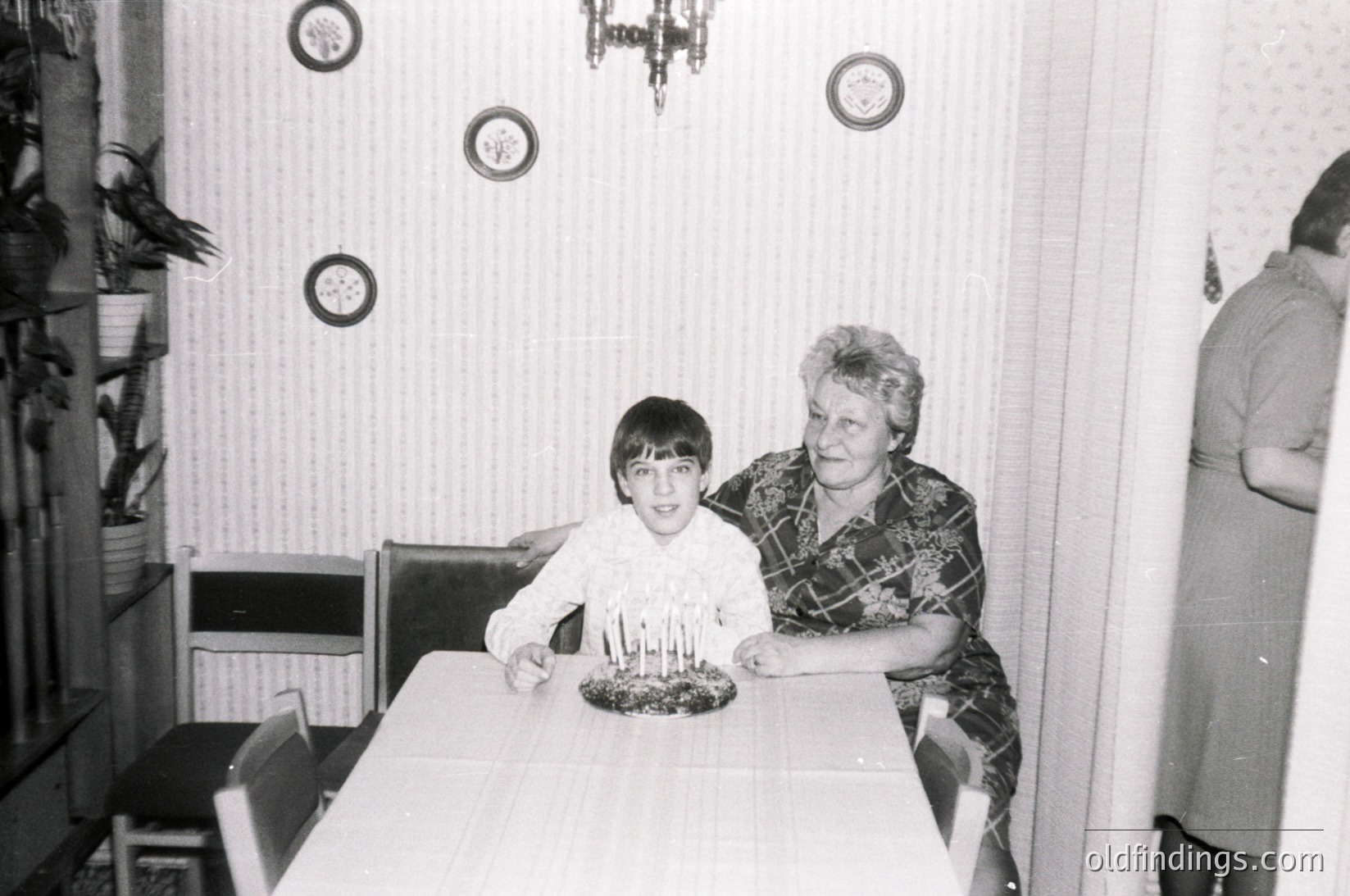A black-and-white indoor family moment: a young boy and an older woman pose at a table with a lit birthday cake. The woman wears a patterned blouse and a headscarf; the boy wears a short-sleeve shirt. Decorative wall clocks and a chandelier-style light fixture add vintage charm. Mid-century interior with simple furnishings.