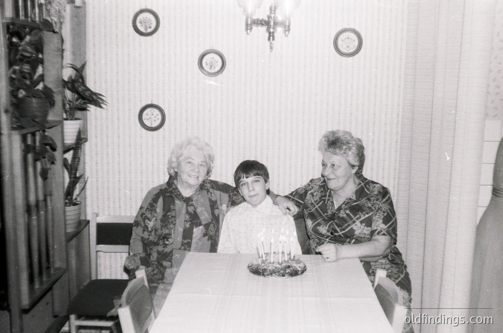 Three generations share a celebratory moment in a modest indoor setting, likely a 1970s–1980s home. A young boy stands between two women at a table adorned with a tiered candle cake. Decorative wall plaques and a shelf with ceramic items frame the scene. Warm familial bond captured in vintage black-and-white.