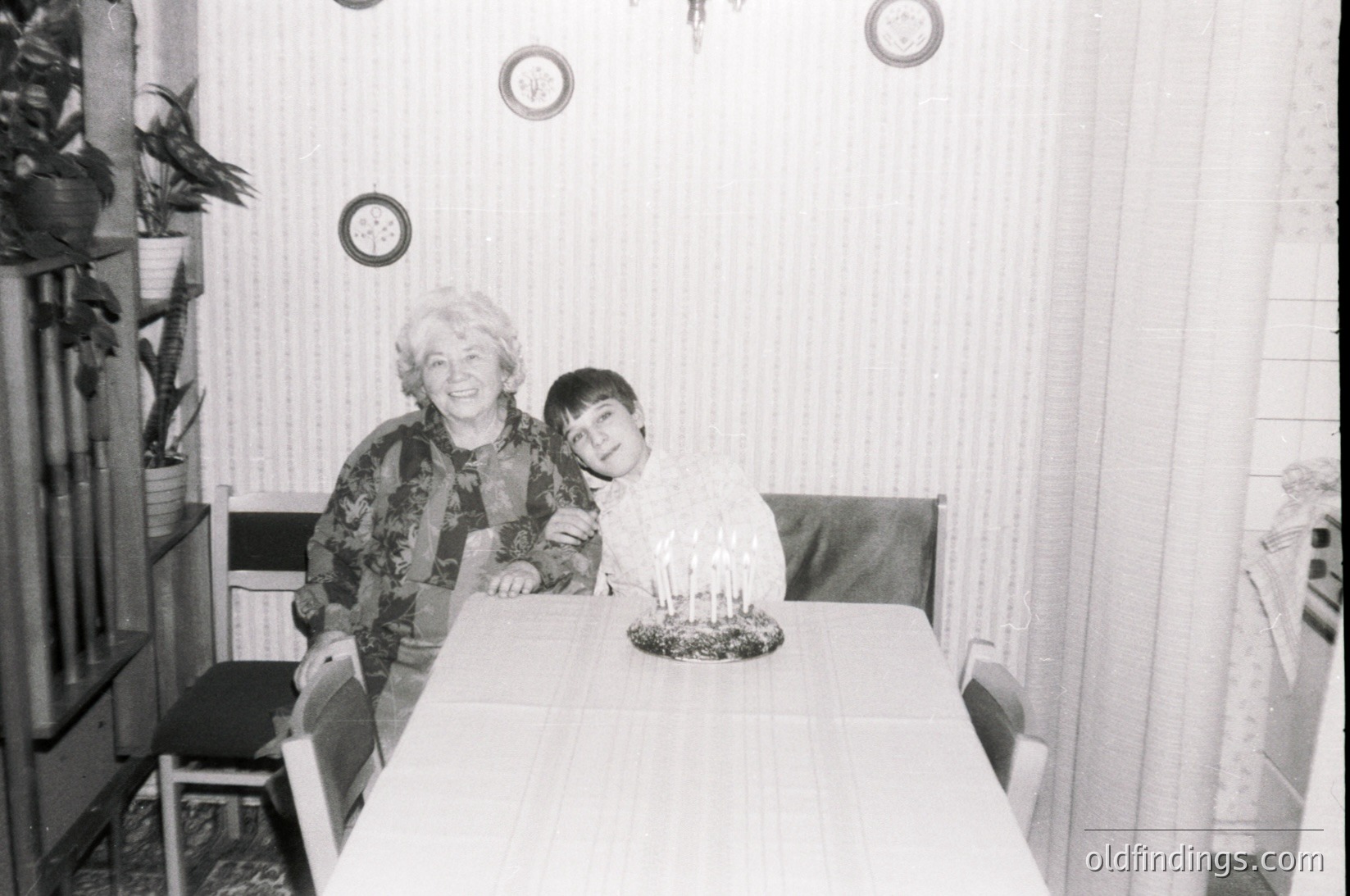 A mid-20th century indoor portrait showing a woman and young boy seated at a simple wooden table. The woman wears patterned clothing, while the boy sits beside a cake with lit candles. Decor includes wall-mounted clocks, a curtained window, and a potted plant. Likely Eastern European, 1960s-1980s.
