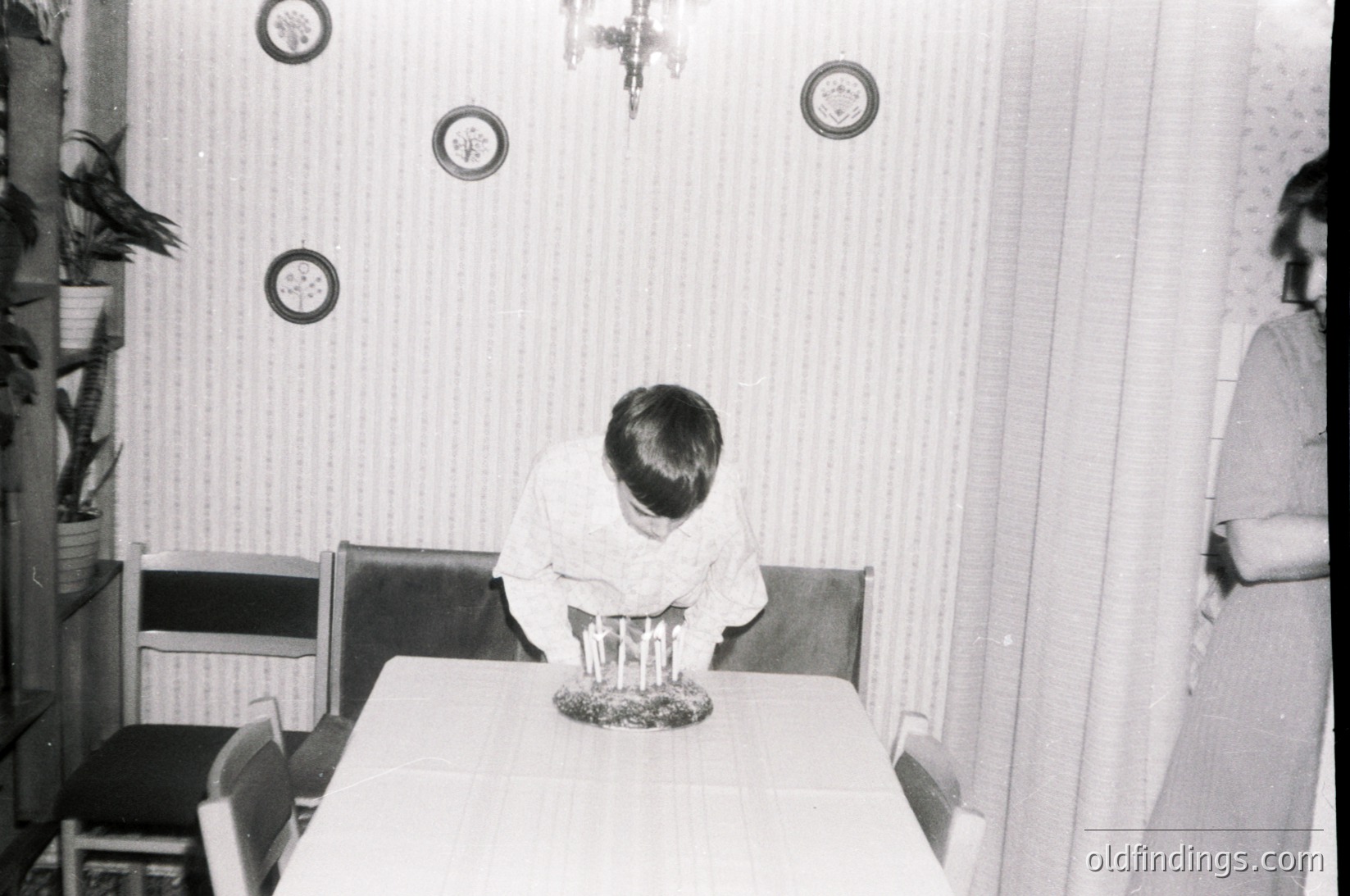 Mid-century dining scene with child seated at round table, back to camera. Plain wallpaper, circular wall decorations, and vintage chandelier visible. Simple wooden chairs and basic kitchen setup suggest 1960s-1970s domestic life.