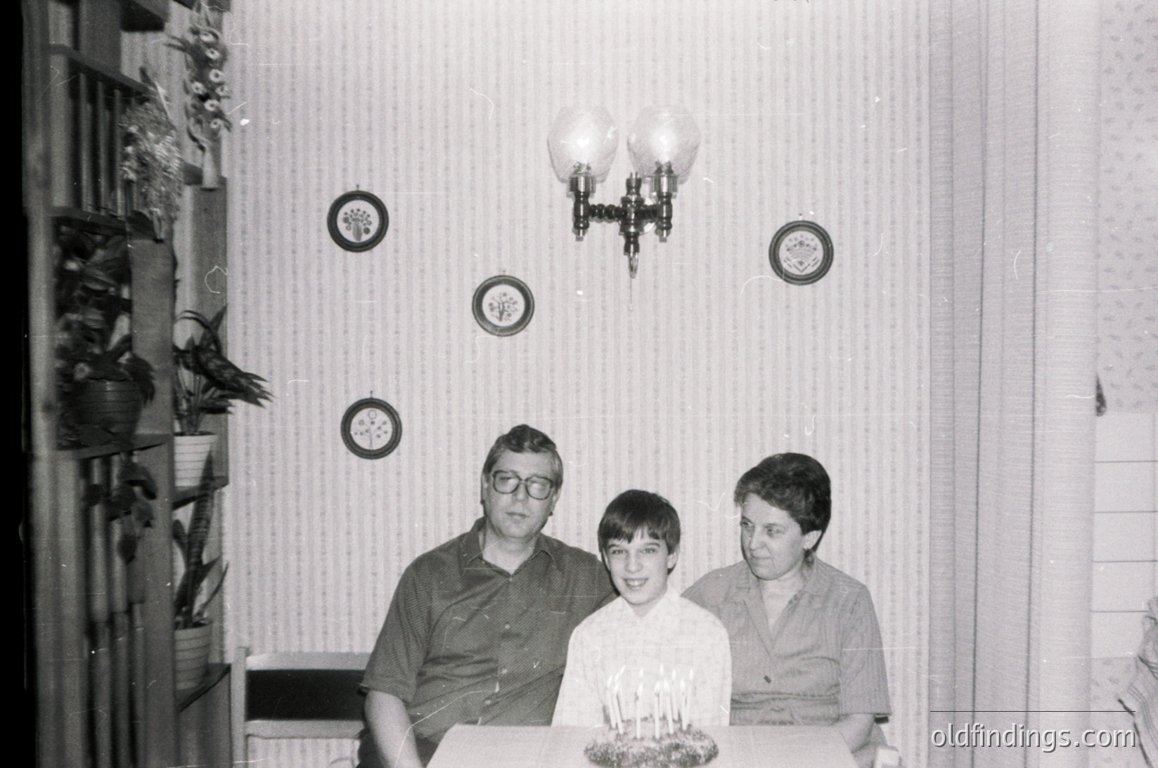 Vintage black-and-white family portrait featuring three generations: a man in a dark shirt, a boy in a white shirt with a lit birthday cake, and a younger boy in a light shirt. Decor includes wall-mounted circular plaques, a chandelier, and a shelf with decorative items. Likely 1960s–1970s domestic setting.