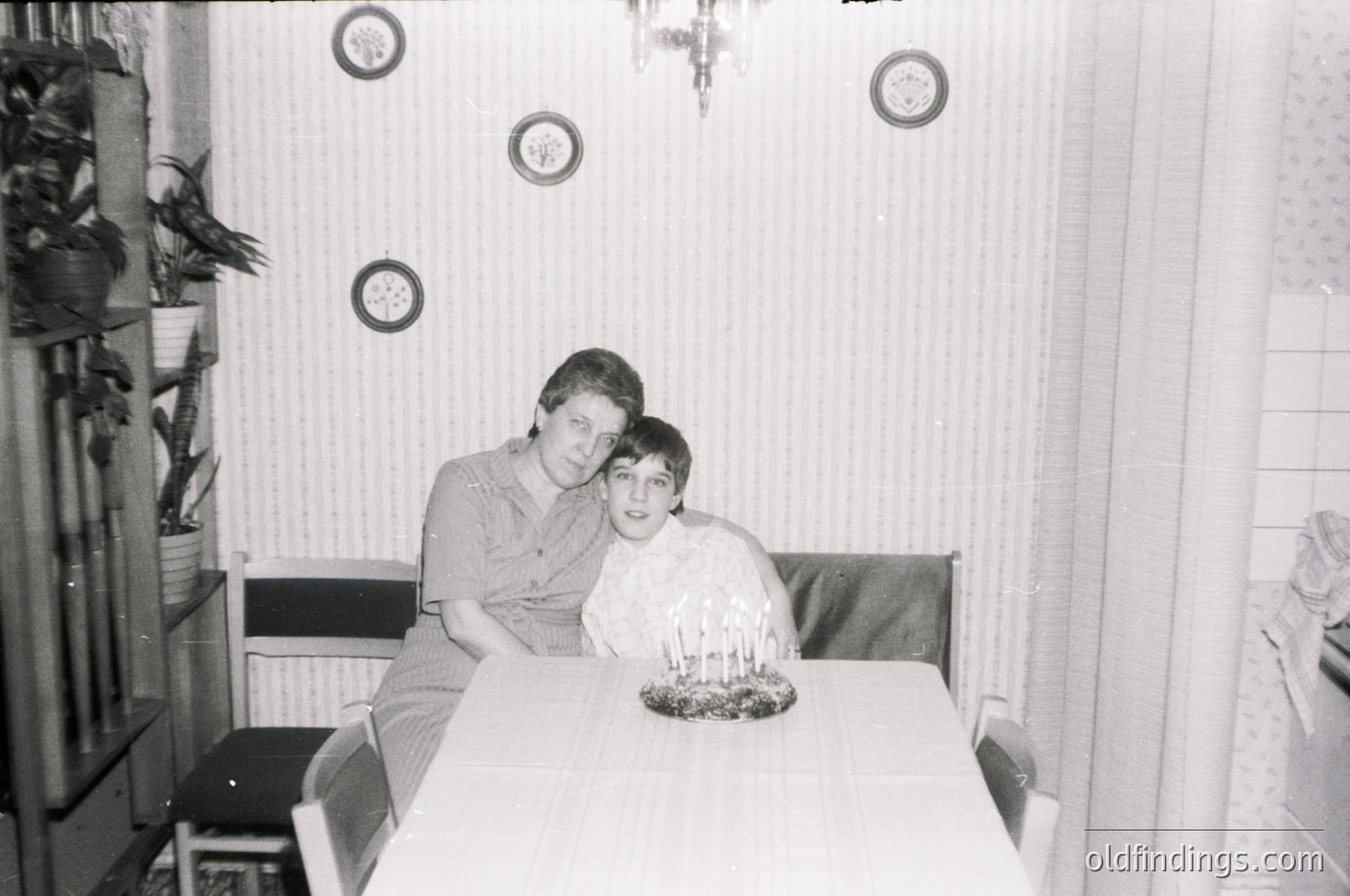 Mid-20th century indoor portrait: two individuals seated at a small round table in a modestly furnished room, likely 1960s–1970s. Circular wall clocks and a potted plant add vintage charm. Formal attire suggests a special occasion or family gathering.