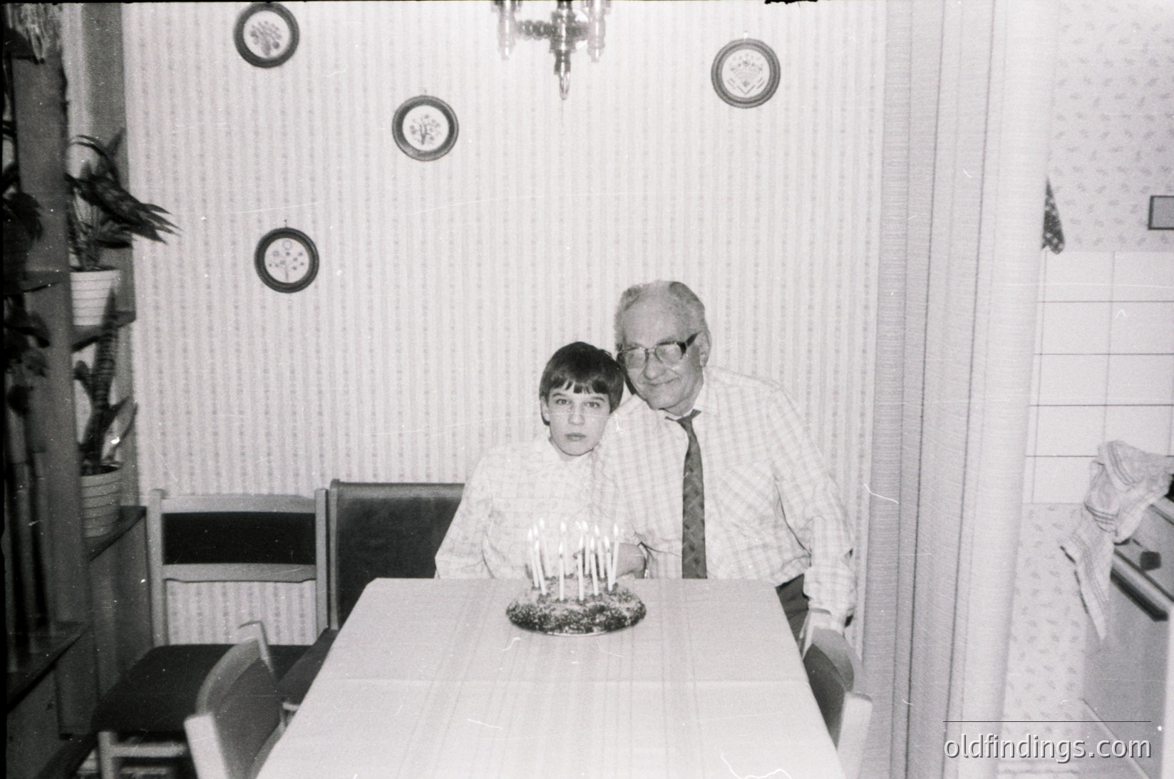 Mid-20th century indoor family portrait: man in suit and tie poses with young boy beside a birthday cake with lit candles. Plain wallpaper, round wall clocks, and simple kitchen table visible. Likely 1960s–1970s domestic setting.