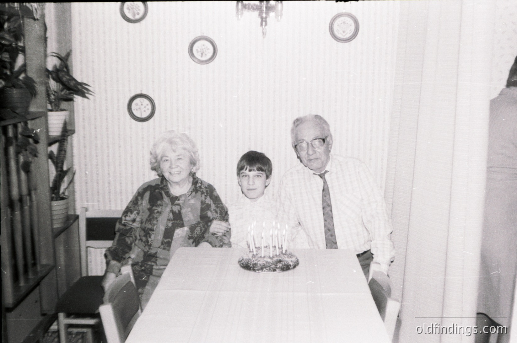 A black-and-white family portrait from the 1970s–1980s, likely Eastern Europe. Three generations seated at a table: an elderly woman in patterned blouse, a young boy in a collared shirt, and an older man in a suit with a tie. Centerpiece features lit candles on a cake. Decor includes wall clocks, a chandelier, and a curtained window.