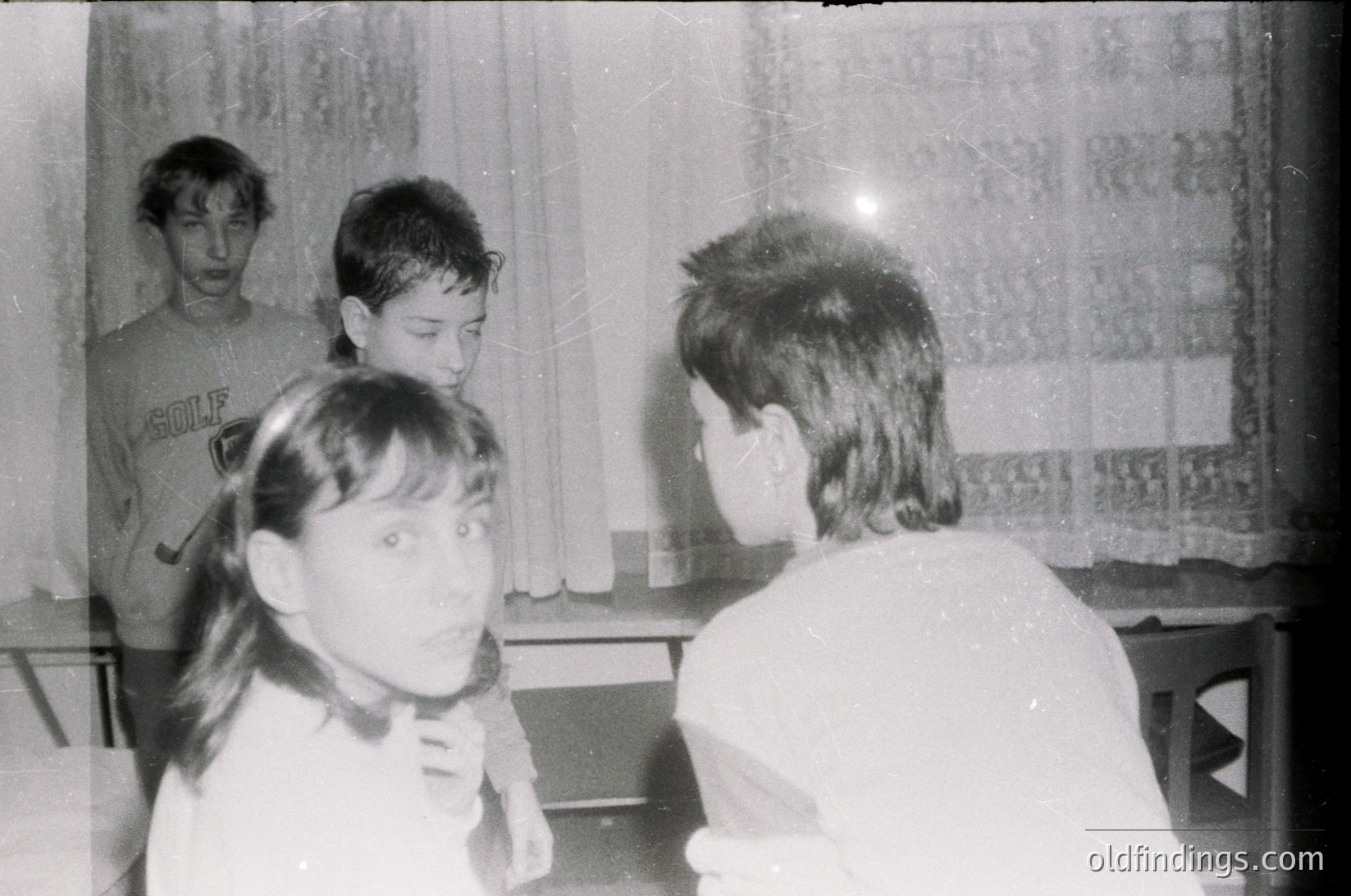 Vintage black-and-white photo of four children in a dimly lit indoor setting, likely a classroom or home. One child wears a "Golf" sweatshirt, others in casual clothing. Blurred background suggests motion or focus on subjects. Evokes mid-20th century (1950s–1970s) school or family life.
