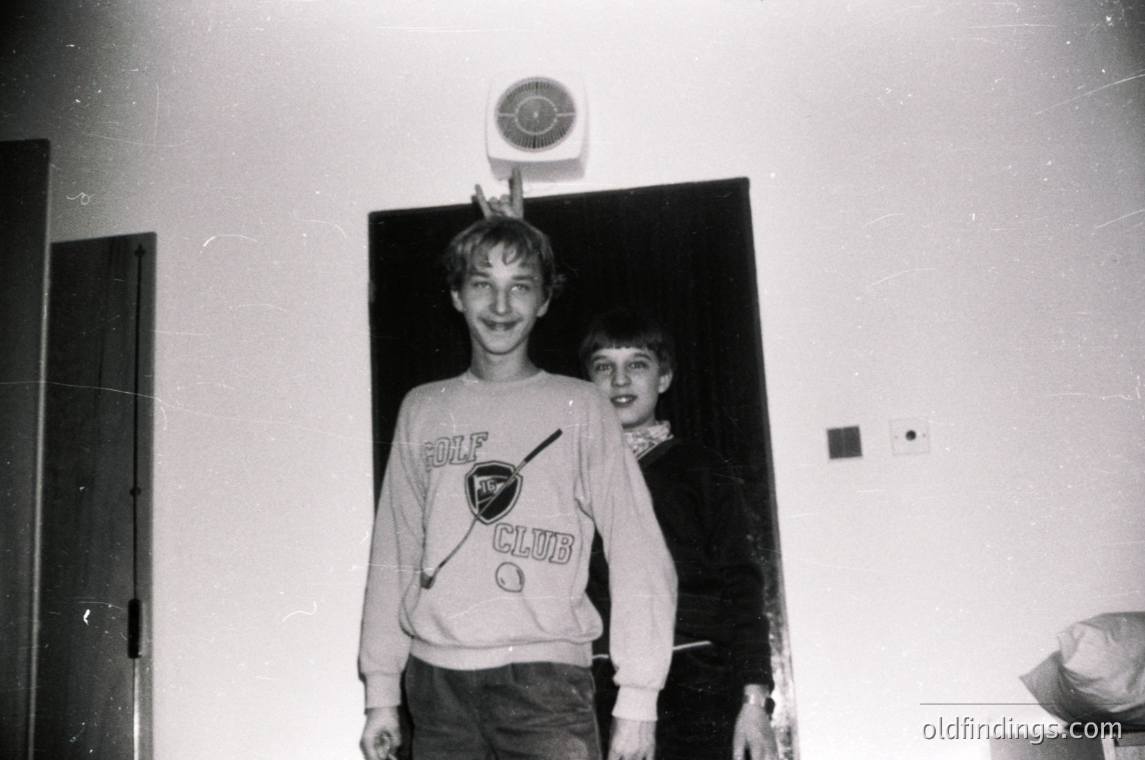 Two boys pose indoors, likely in a 1970s-1980s setting. The boy on left wears a "Golf Club" sweatshirt with a logo, while the other wears a plain sweater. A wall clock and wall socket are visible, suggesting a residential or clubhouse interior.