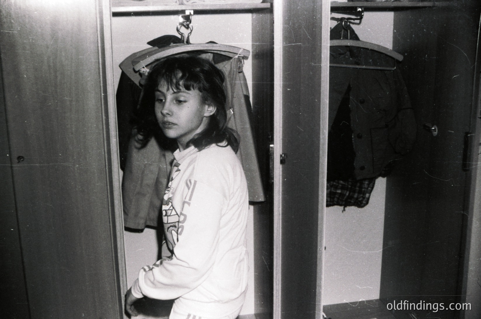 Black-and-white shot of a young individual in a narrow closet, holding a hat above their head. The person wears a long-sleeve shirt with a geometric design and light-colored pants. Closet features metal shelving and hanging garments. Style suggests mid-20th century domestic interior, likely Western Europe or North America.