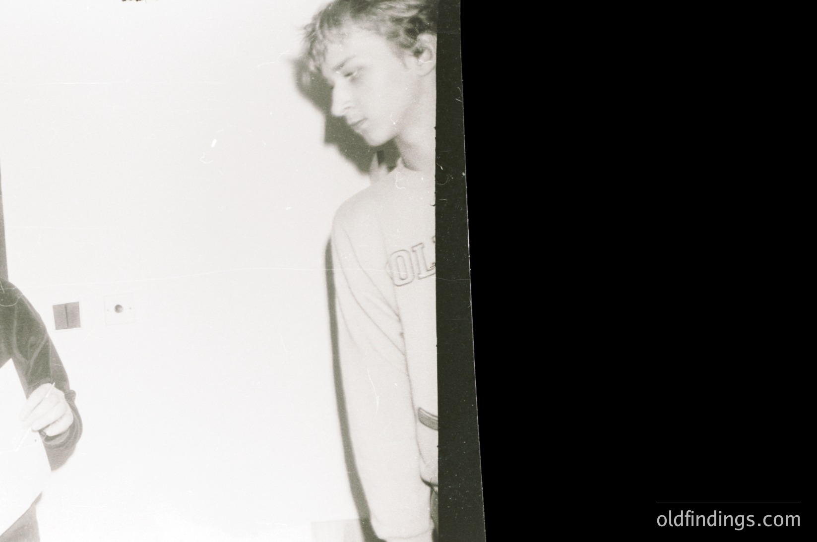 Vintage black-and-white photo of two individuals in a dimly lit indoor setting, likely a 1970s–1980s home or studio. One person wears a sweatshirt with a visible logo, standing near a dark vertical surface. Grainy, underexposed film grain enhances nostalgic aesthetic. Potential use for retro fashion, documentary, or cultural studies research.