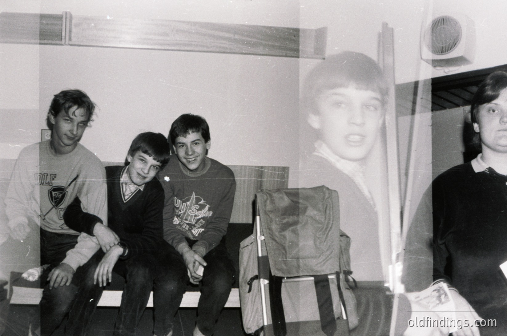 Four young men pose indoors, likely a school or dormitory setting, mid-1970s. Left to right: one wears a sweatshirt with a logo, another sports a sweater with a graphic print, the third has a dark sweater with a logo, and the fourth stands partially visible. A backpack rests beside them. Minimalist interior with a vent and window frame visible.