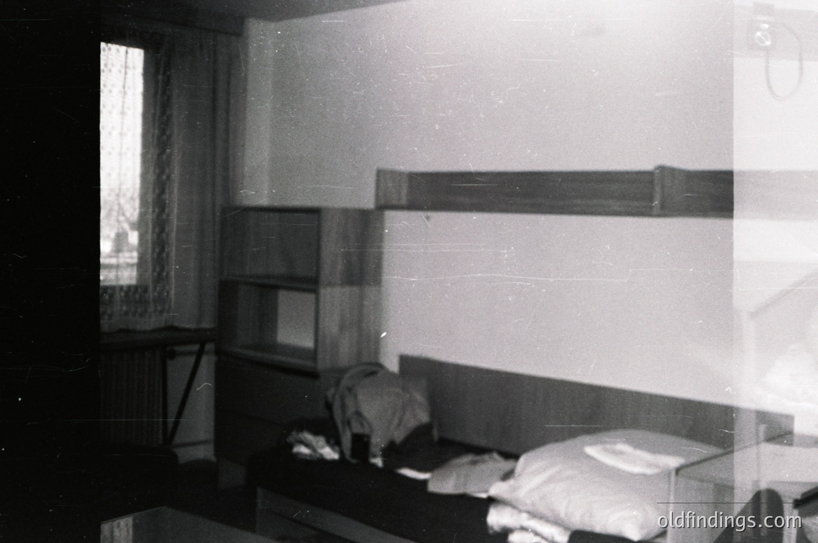 Vintage mid-century bunk bed setup in a minimalist bedroom, featuring built-in shelves above and a single bed below. Plain walls and a window with sheer curtains suggest functional, utilitarian design. Likely residential, possibly a dorm or small apartment.
