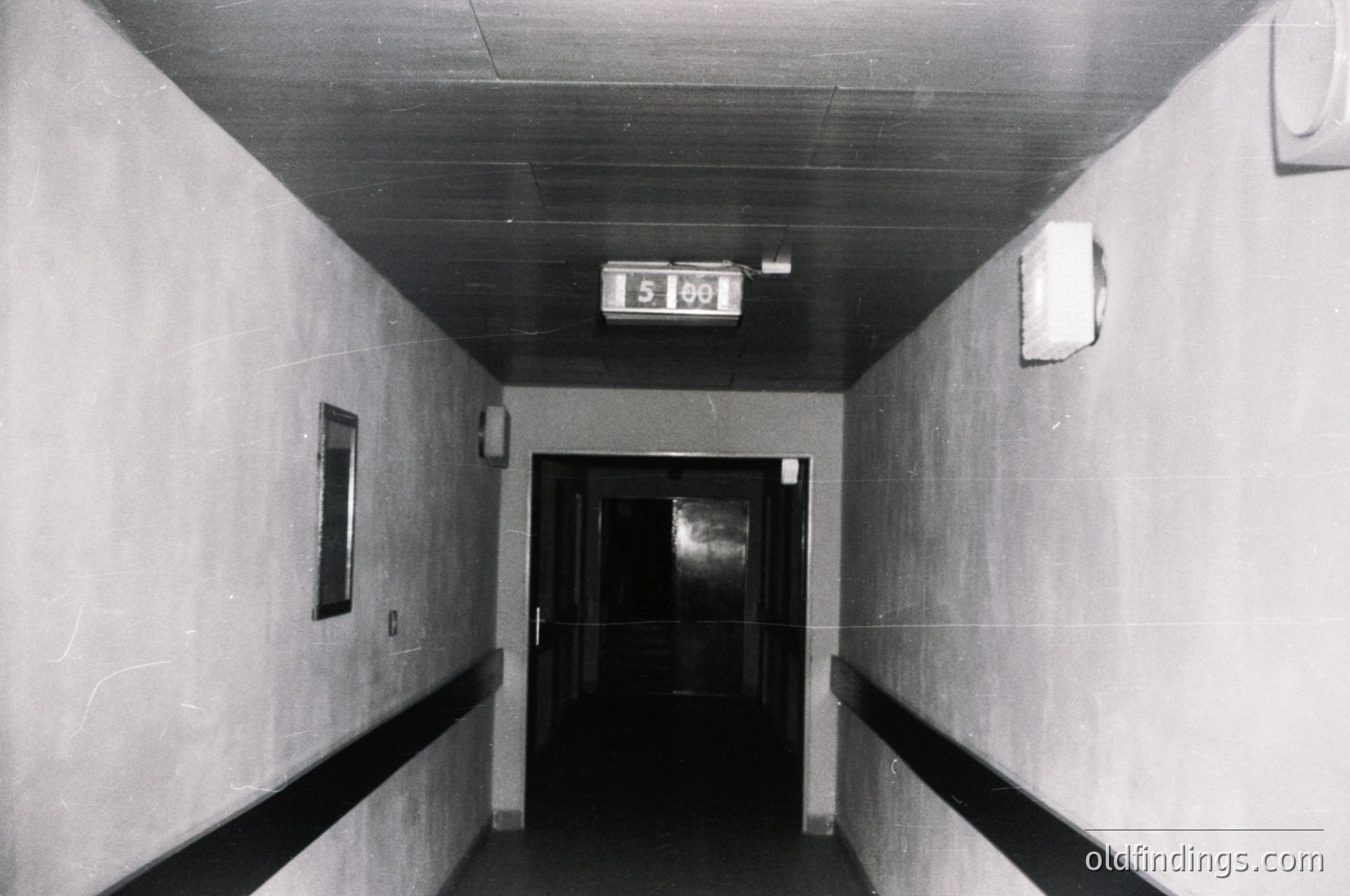 Mid-century institutional hallway with concrete walls and ceiling, featuring a digital floor clock displaying "5:00." Plain door at end with adjacent wall-mounted light fixture. Minimalist, utilitarian design suggests a public or office building from the 1960s–1980s.