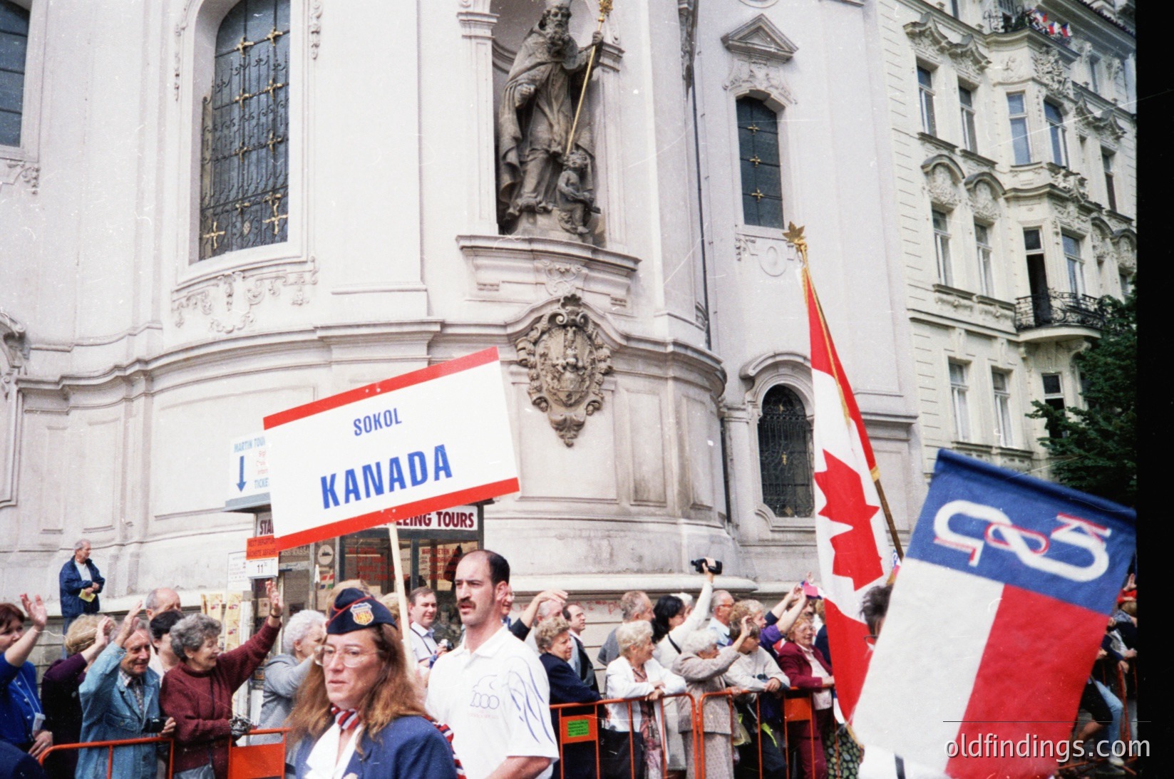 Neoclassical building facade with Baroque statues flanking entrance, crowded with people holding signs and flags. Prominent "SOKOL KANADA" banner and Canadian flags indicate a cultural or heritage event, likely a parade or rally. Mid-20th century urban setting, possibly European.