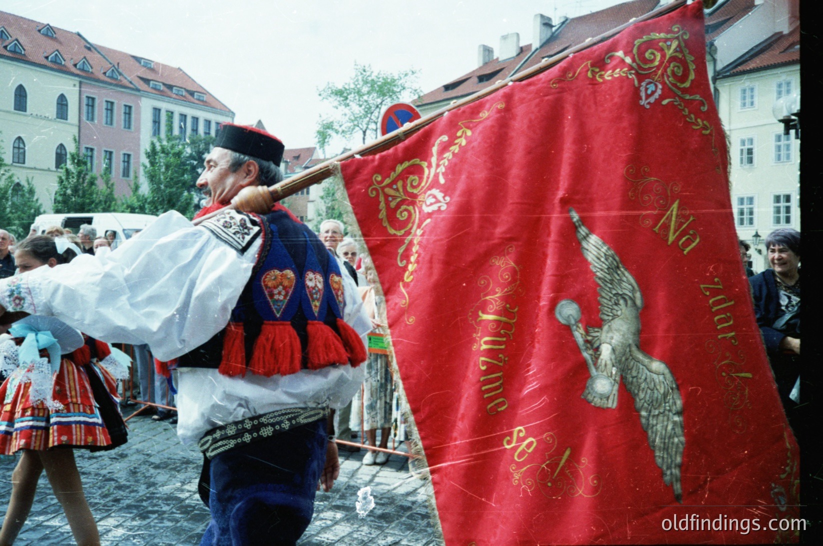 Traditional folk festival participant in ornate attire, carrying a red banner with gold embroidery and a stylized eagle motif. Cobblestone street and historic European architecture in background. Likely Eastern European cultural celebration, mid-20th century.