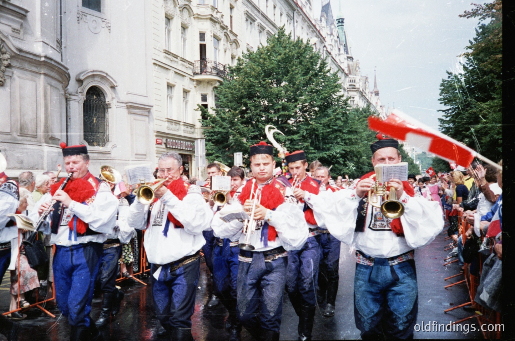 Uniformed brass band performing in a European city street, likely 1980s–1990s. Members wear white jackets with red accents, blue trousers, and red hats with white trim. Crowd and historic architecture in background suggest a festive public event. Ideal for historical research or cultural documentation.