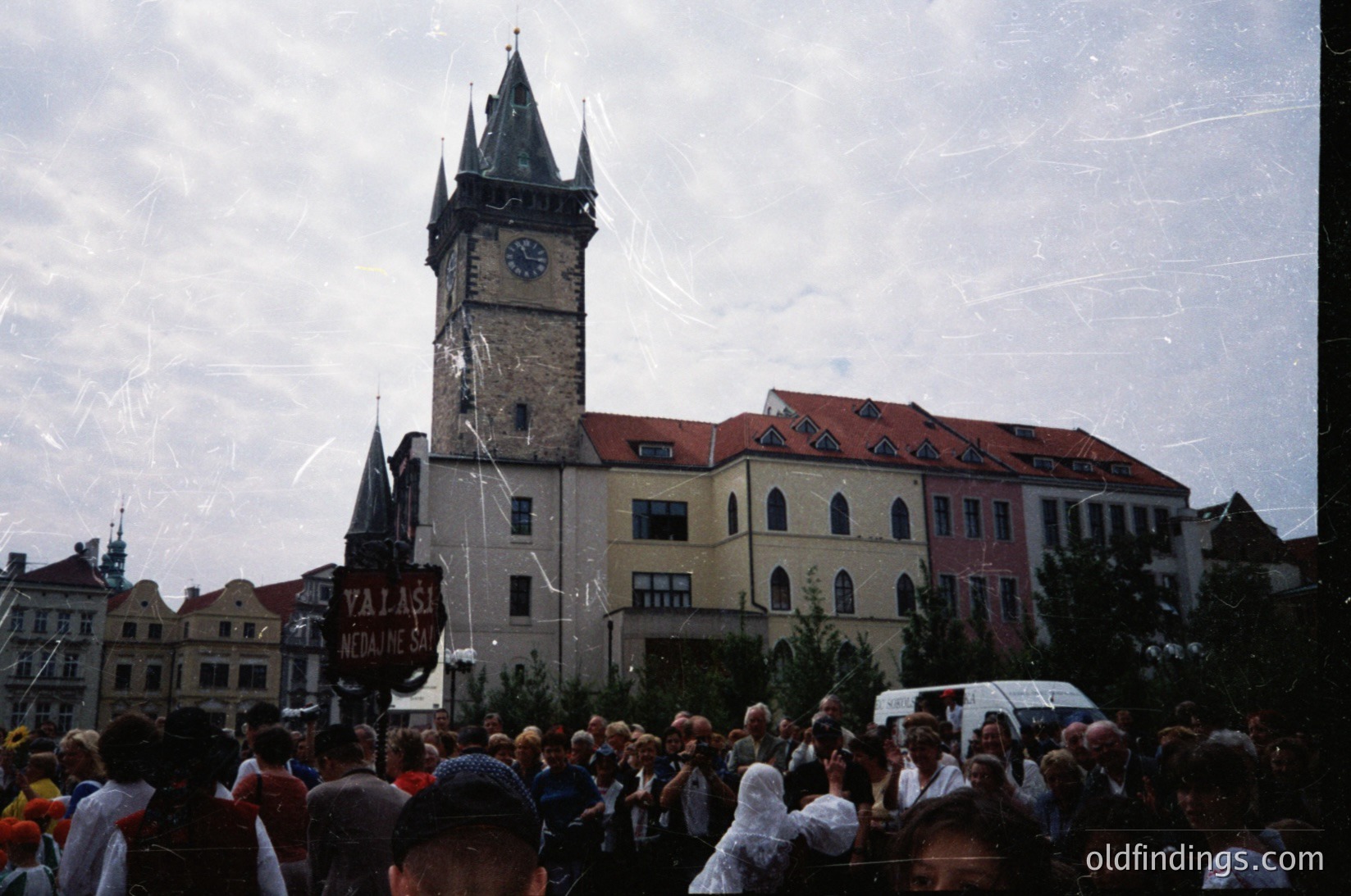 Historic clock tower with Gothic Revival architecture dominates a crowded public square. Crowd gathered beneath a banner reading "Valašské Meziříčí" in Czech, likely during a festival or market. Red-tiled roofs and cream-colored buildings frame the scene. Colorful, vintage filter enhances warm tones. šskéMeziříčí