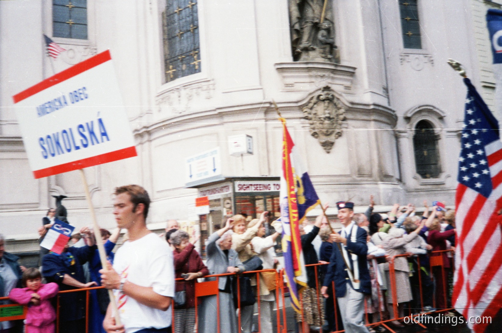 Crowd rally near grand neoclassical building with "Sightseeing Tour" signage. Participants hold flags: American, Polish, and "Ameryka Obc" Sokolska banner. Formal attire suggests 1960s–1970s era. Prominent statue atop building. Crowd gathered behind barriers, likely a cultural or patriotic event.